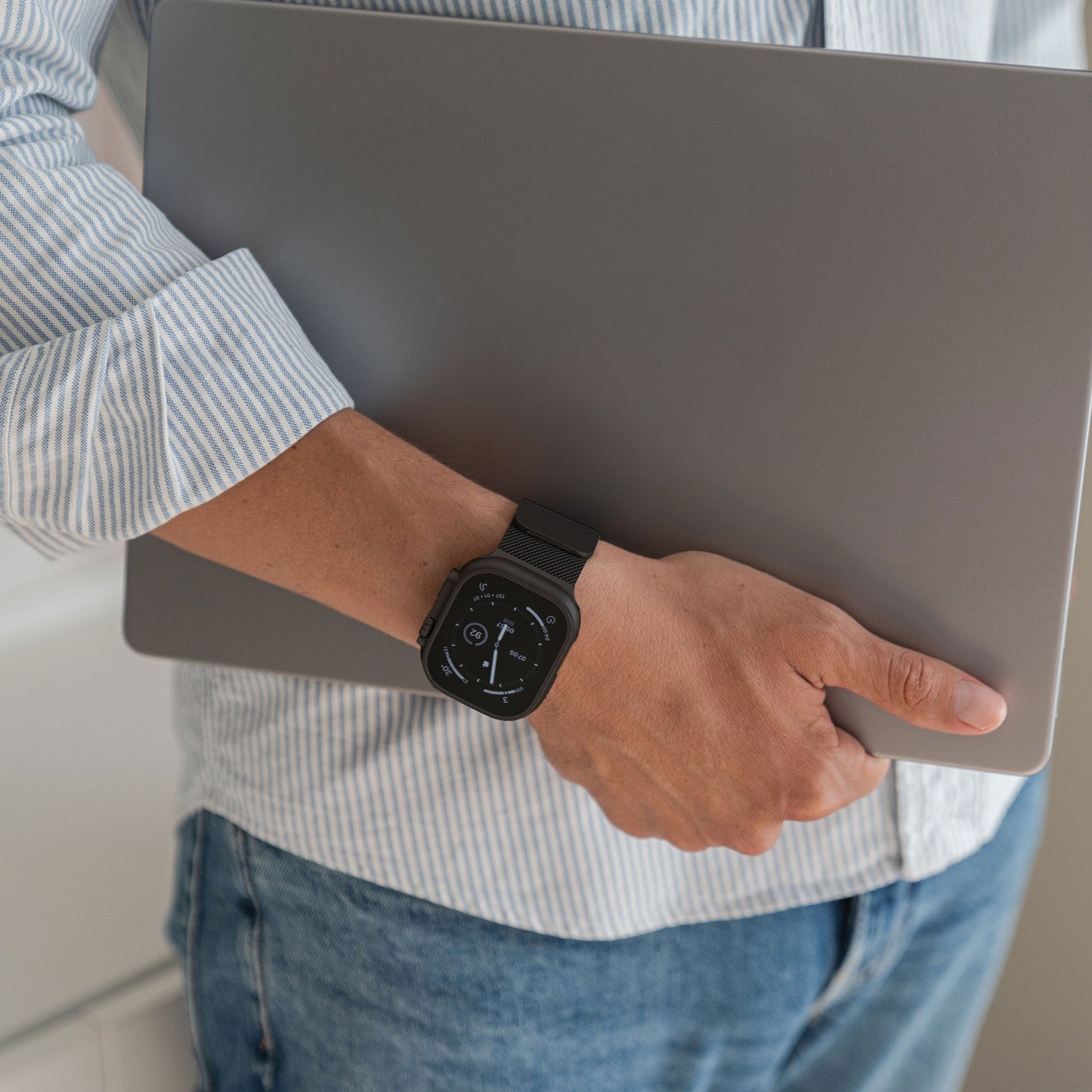 Eine Person in einem gestreiften Hemd hält einen geschlossenen Laptop und trägt das arktisband Apple Watch Ultra Milanaise Armband von arktisband am Handgelenk.