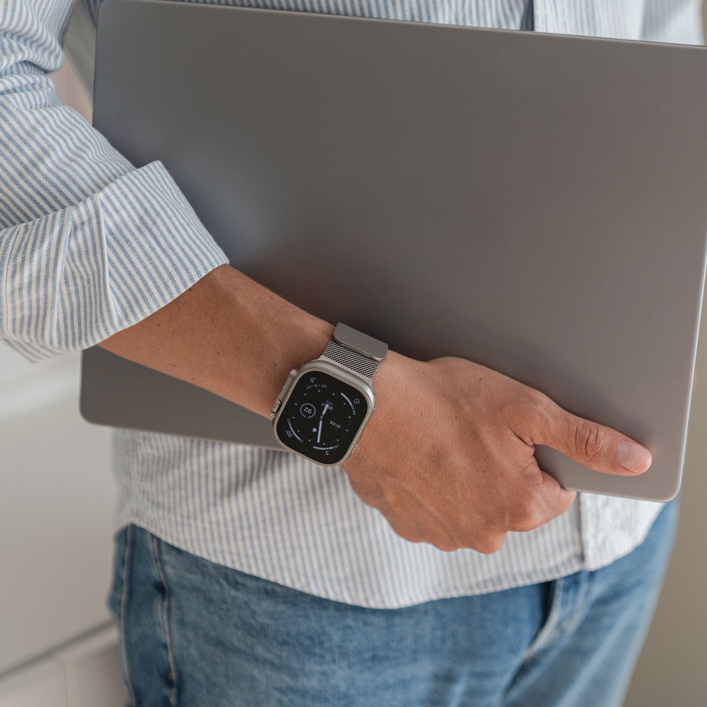 Eine Person in einem gestreiften Hemd hält einen geschlossenen Laptop und trägt das arktisband Apple Watch Ultra Milanaise Armband von arktisband am Handgelenk.