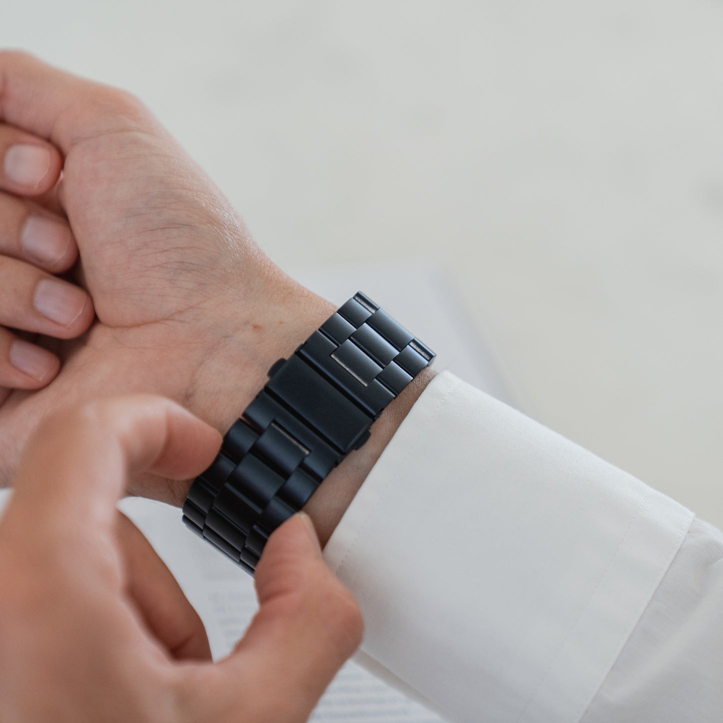 Eine Person in einem weißen Langarmshirt rückt ein Arktisband Apple Watch Gliederarmband „Arctica“ an ihrem linken Handgelenk zurecht, im Hintergrund ist ein unscharfes Dokument zu sehen.