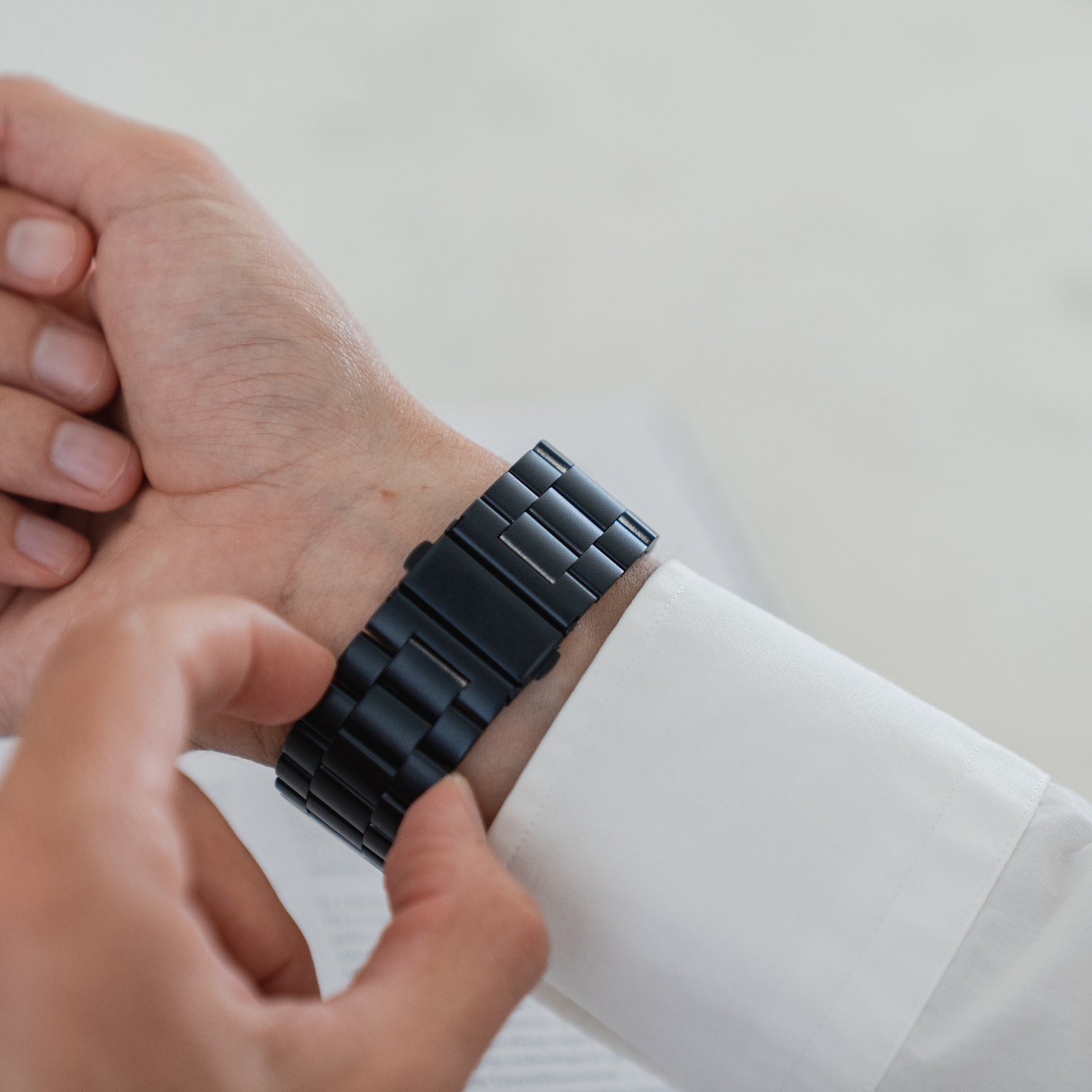 Eine Person in einem weißen Langarmshirt rückt ein Arktisband Apple Watch Gliederarmband „Arctica“ an ihrem linken Handgelenk zurecht, im Hintergrund ist ein unscharfes Dokument zu sehen.