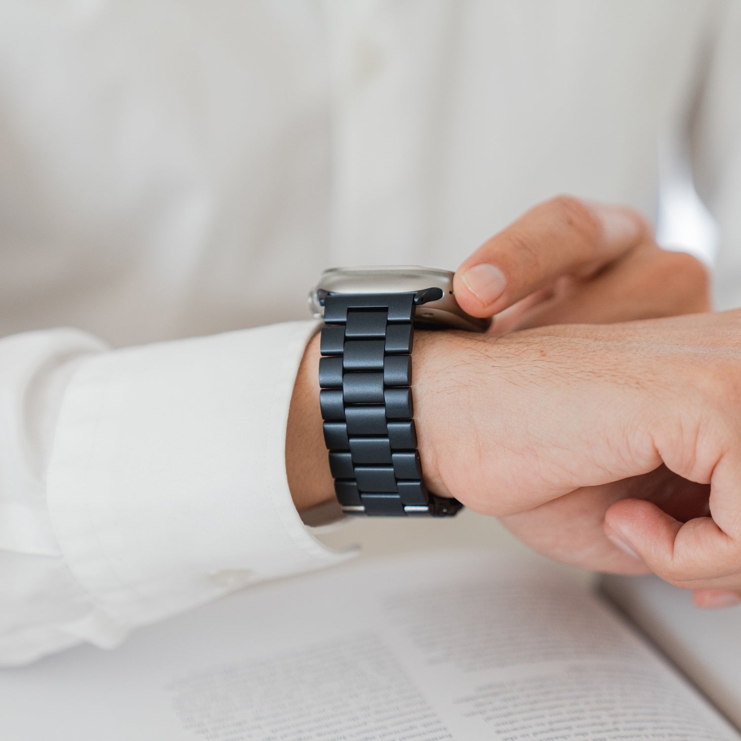 Eine Person im weißen Hemd checkt ihre Smartwatch mit dem Arktisband Apple Watch Gliederarmband „Arctica“ in Schwarz, vor ihr liegt ein aufgeschlagenes Buch auf dem Tisch.