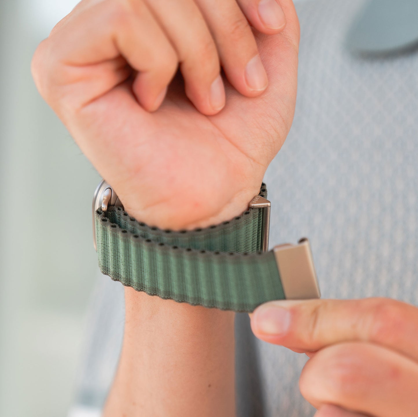 Jemand befestigt das grüne arktisband Apple Watch Nylonarmband „Venture“ am Handgelenk und schließt den Verschluss. Der stylische Wave-Look macht dieses arktisband perfekt für Outdoor-Fans – abgebildet vor einem sanft verschwommenen Hintergrund.
