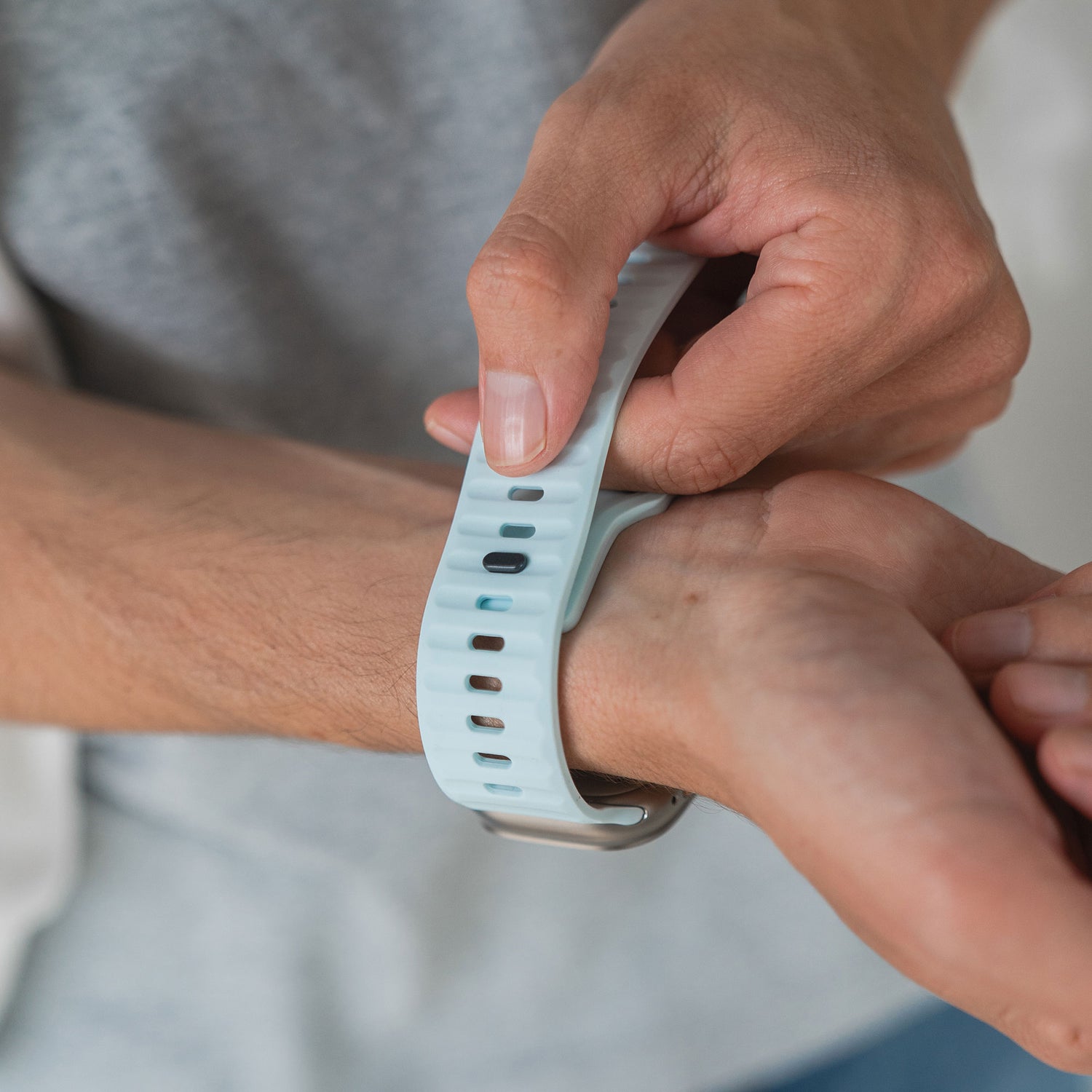 Jemand befestigt das arktisband Apple Watch Silikonarmband „Active“ in Hellblau am Handgelenk, beide Hände sind sichtbar, und trägt ein graues Shirt für einen sportlichen Look.