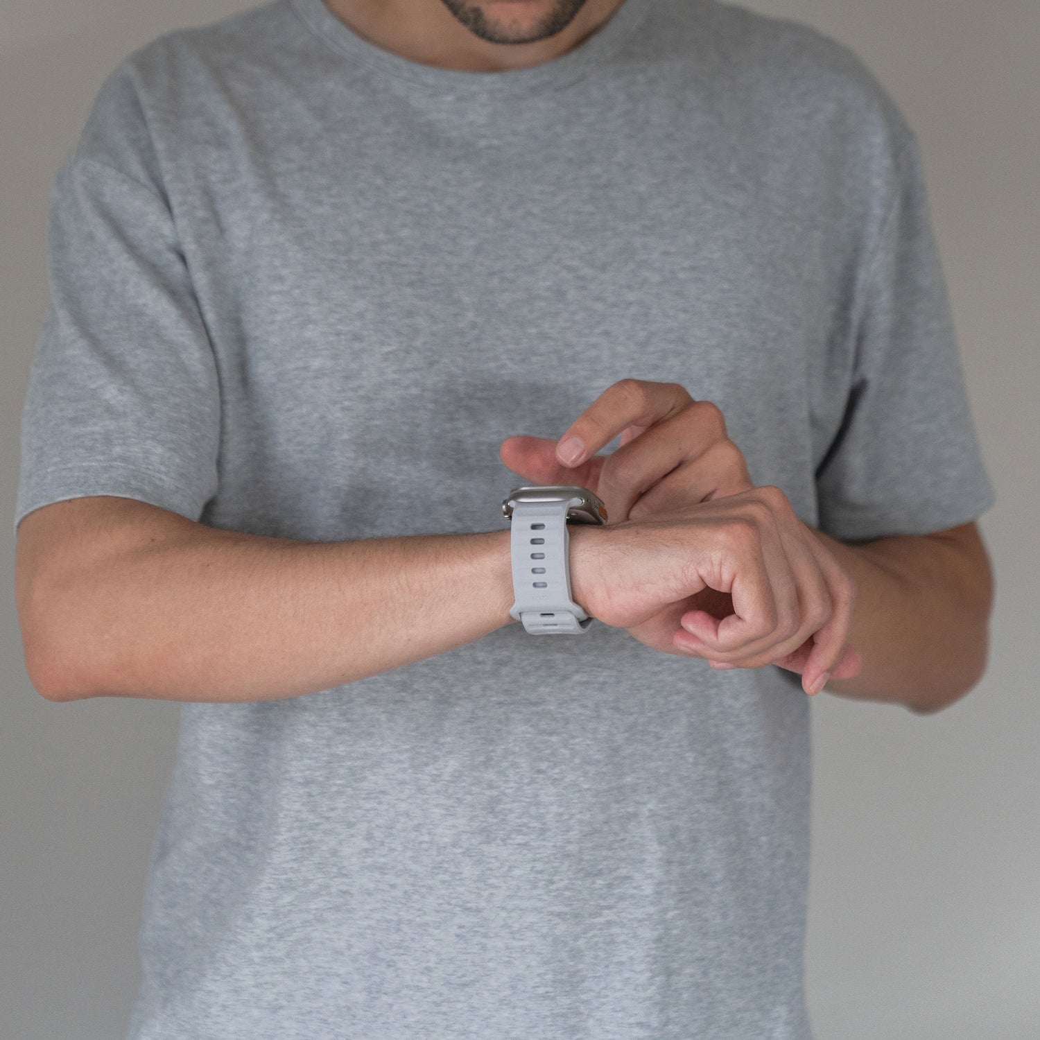Eine Person in einem grauen T-Shirt steht vor einem schlichten Hintergrund und interagiert mit ihrer rechten Hand mit einem arktisband Apple Watch Silikonarmband „Active“ von arktisband an ihrem linken Handgelenk für einen sportlicheren Look.