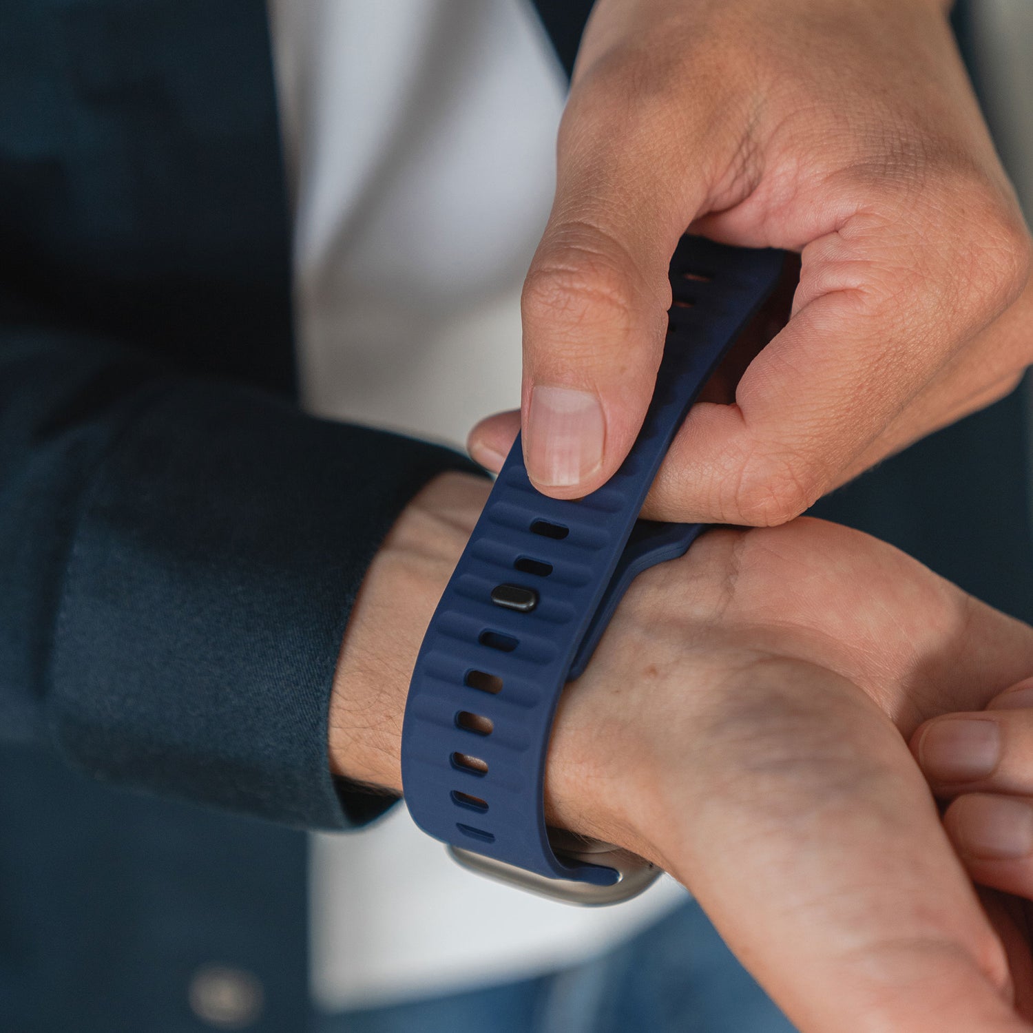 Eine Person befestigt das arktisband Apple Watch Silikonarmband „Active“ in Blau mit beiden Händen am Handgelenk und trägt für einen sportlichen Look eine dunkle Jacke und ein weißes Hemd.