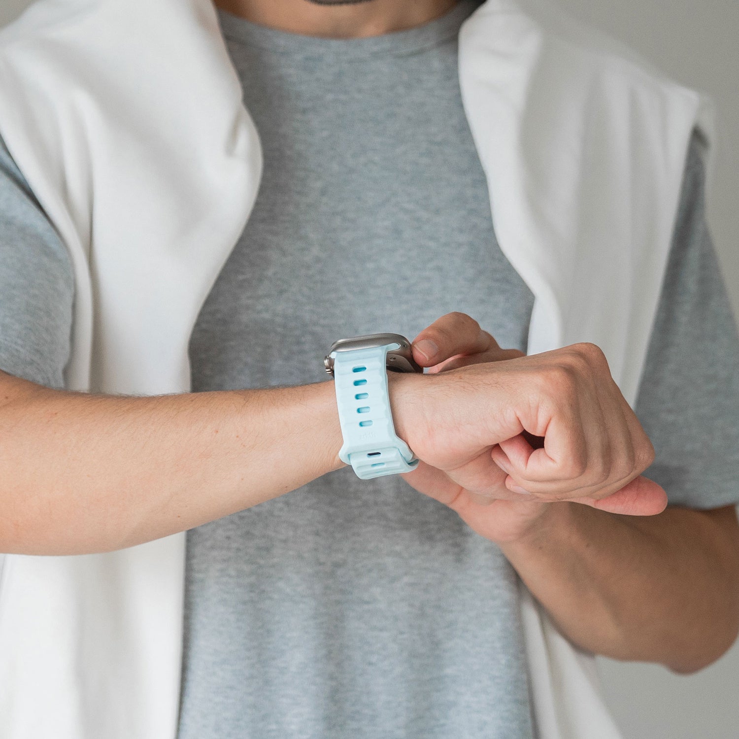Eine Person in einem hellgrauen T-Shirt und einem weißen Pullover checkt ihre Apple Watch. Das arktisband Apple Watch Silikonarmband „Active“ von arktisband in Hellblau sorgt vor einem neutralen Hintergrund für einen sportlicheren Look.
