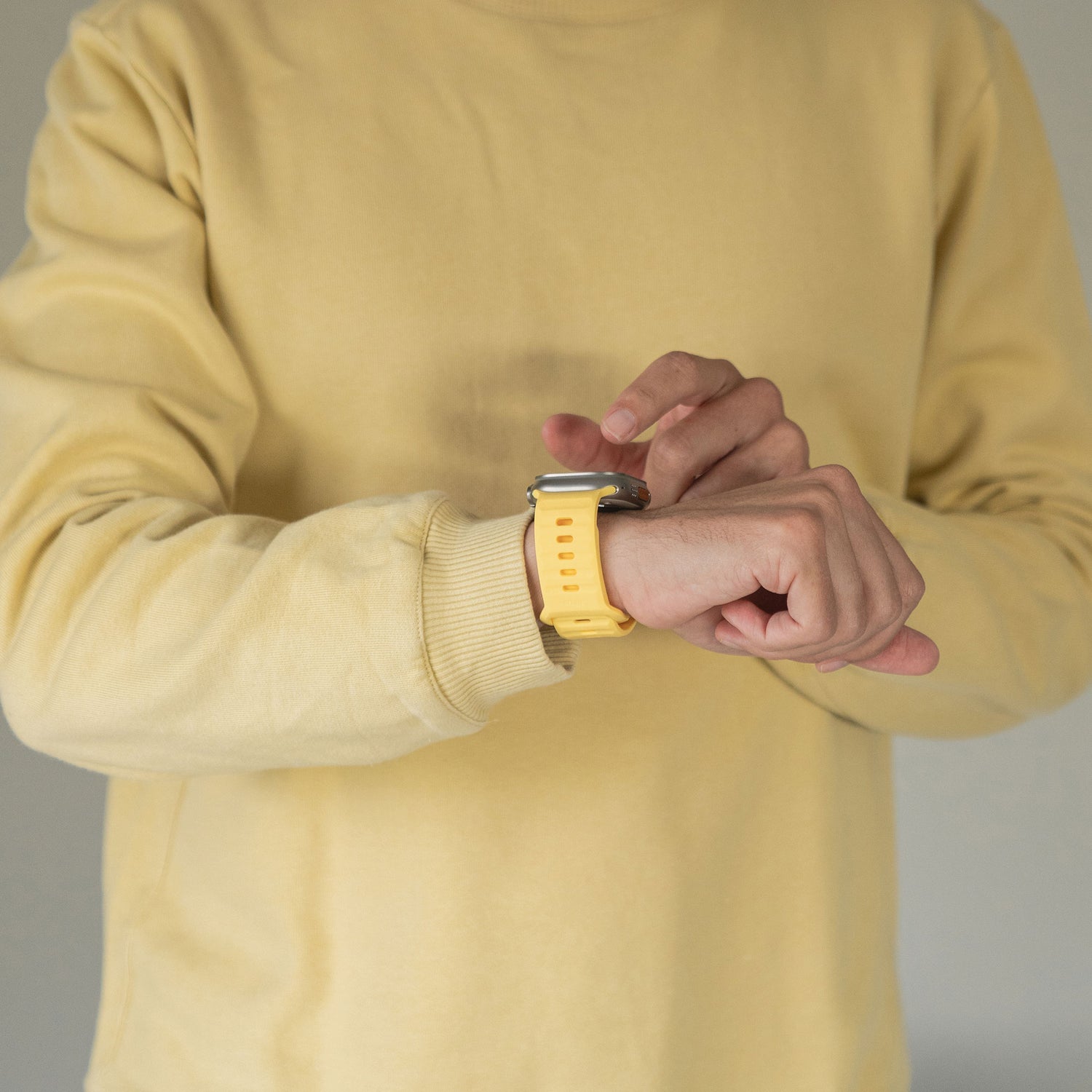 Eine Person im gelben Pullover rückt mit der rechten Hand ihr arktisband Apple Watch Silikonarmband „Active“ von arktisband, das sie am linken Handgelenk trägt, zurecht. Der Hintergrund ist schlicht und neutral.