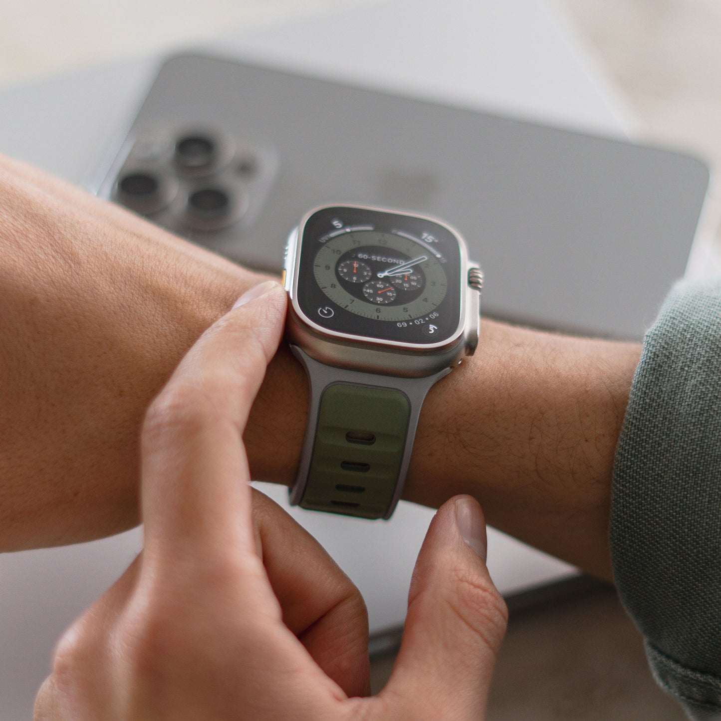 Eine Person, die ein Arktisband Apple Watch Silikonarmband „Swift“ mit grünem Band und Aluminium-Pin-Verschluss trägt, hält ihr Handgelenk in die Nähe eines Smartphones und eines Laptops.