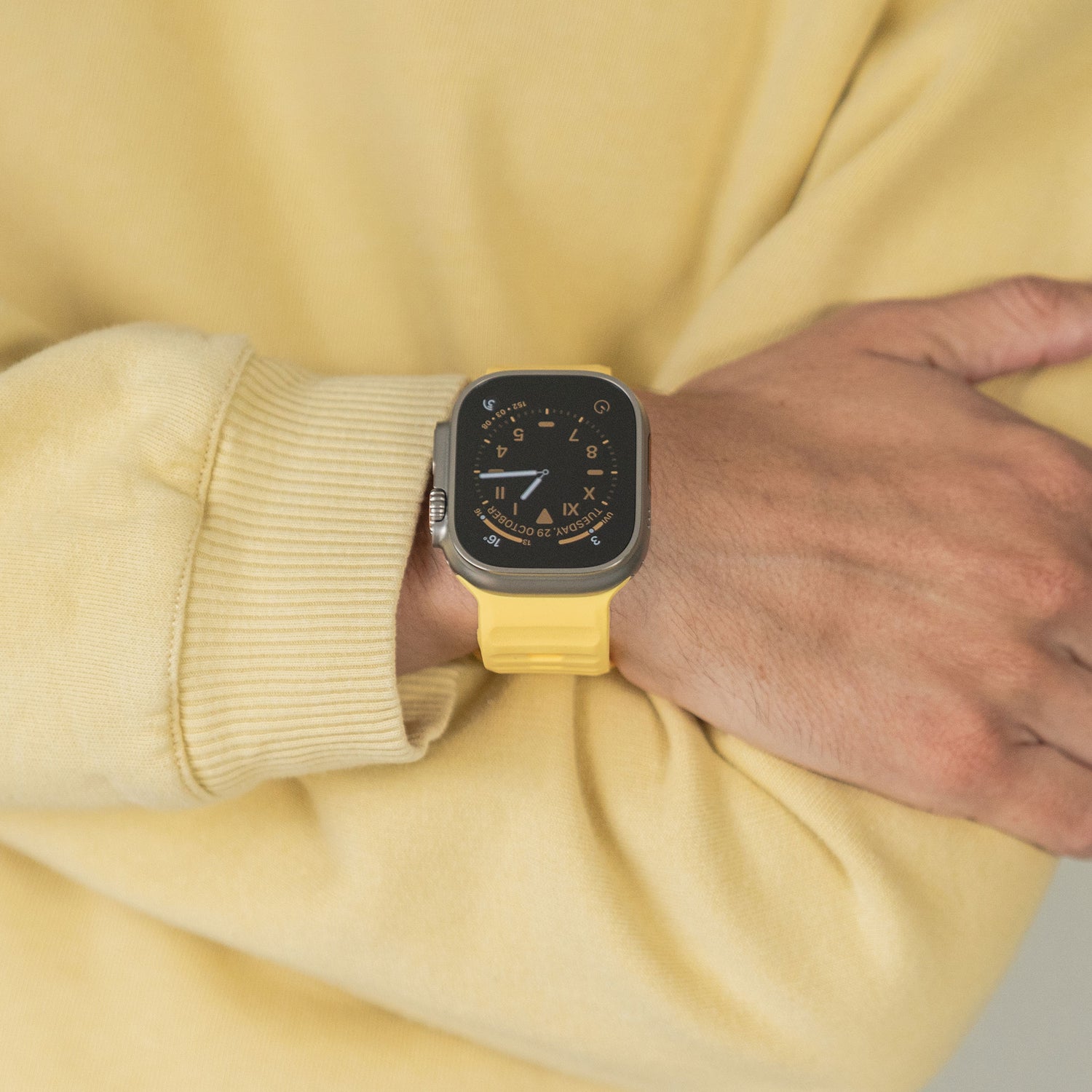 Eine Person, die ein gelbes Sweatshirt und ein dazu passendes gelbes Arktisband Apple Watch Silikonarmband „Active“ mit schwarzem rechteckigem Zifferblatt trägt und für einen sportlichen Look den rechten Arm vor dem Körper verschränkt.
