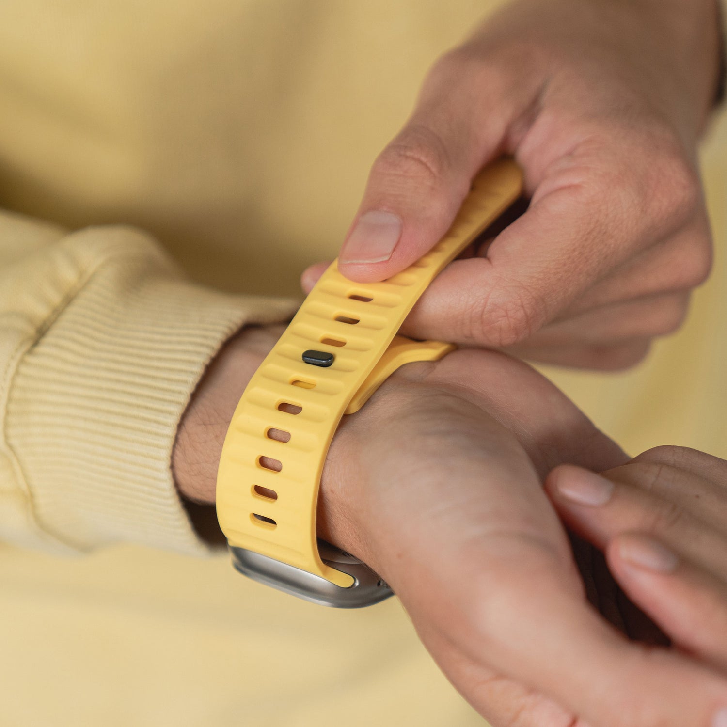 Eine Person mit einem gelben Langarmshirt befestigt das arktisband Apple Watch Silikonarmband „Active“ von arktisband um ihr Handgelenk und passt es für einen sportlichen Look und sicheren Sitz an.