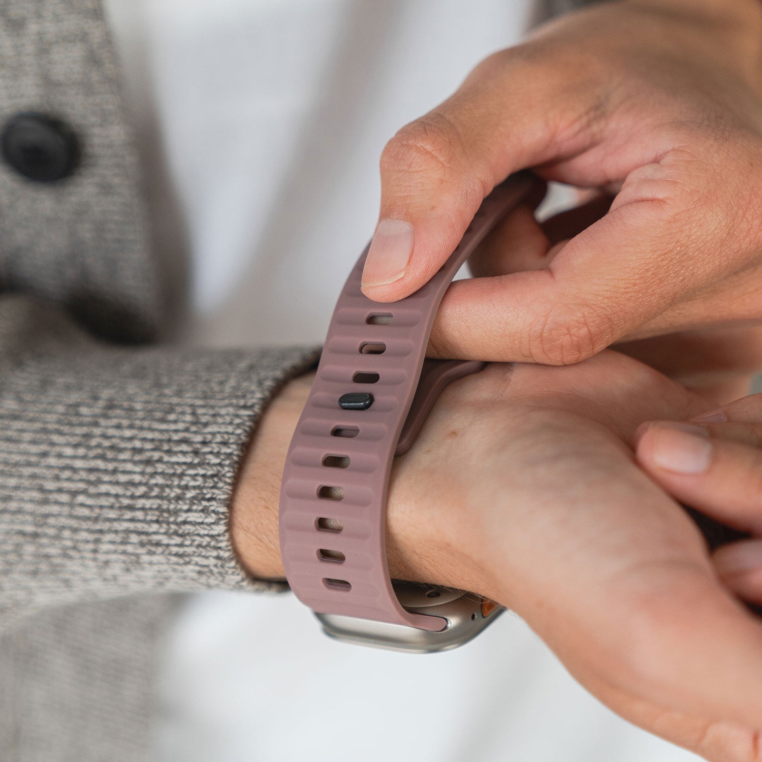 Eine Person befestigt das arktisband Apple Watch Silikonarmband „Active“ in Mauve und nutzt die vielen Löcher für einen sportlichen Look. Dazu trägt sie einen grauen Strukturpullover.