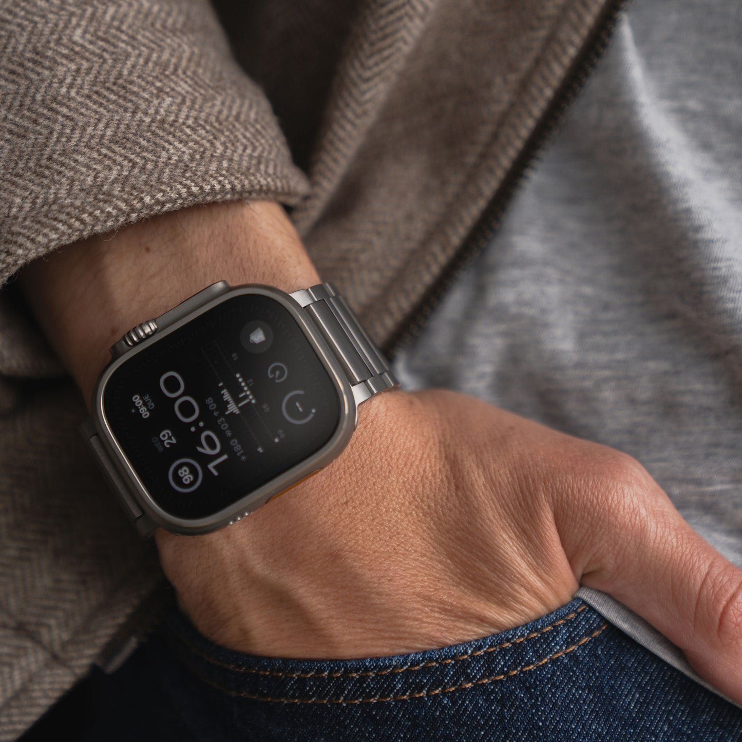 Eine Person trägt ein Arktisband Apple Watch Ultra Titanarmband „Maverick“ am linken Handgelenk, dazu ein graues Hemd, eine braune Fischgrätjacke und blaue Jeans. Die Hand in der Tasche. Die Uhr zeigt 16:00 Uhr.