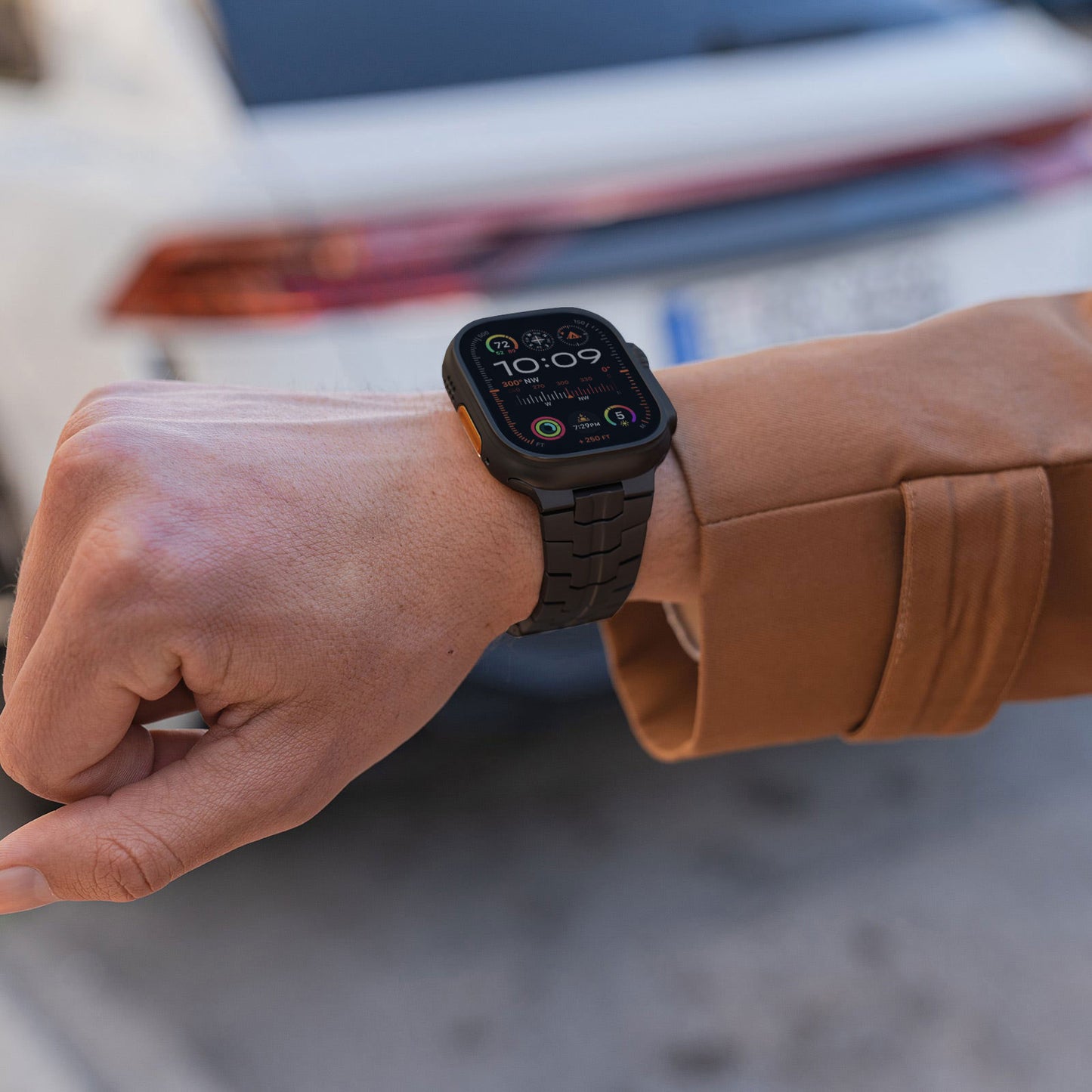 Eine Person in einer braunen Jacke zeigt an ihrem linken Handgelenk ein arktisband Apple Watch TITAN Armband „Aviato Edition“ von arktisband, das 10:09 anzeigt, während sie draußen neben einem verschwommenen weißen Auto steht.