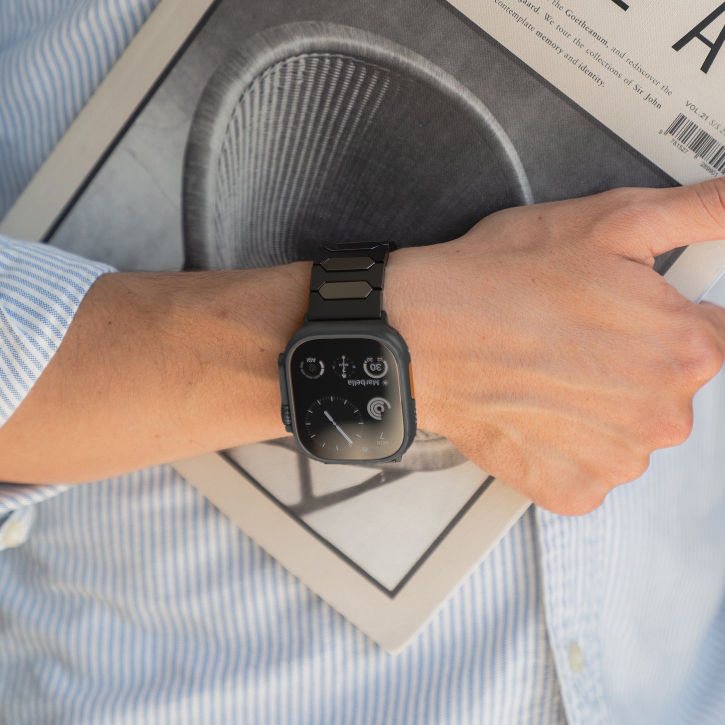 Eine Person, die ein arktisband Apple Watch Ultra Titanarmband "Vectoron" von arktisband trägt, hält eine Zeitschrift mit einem kreisförmigen Design auf dem Cover.