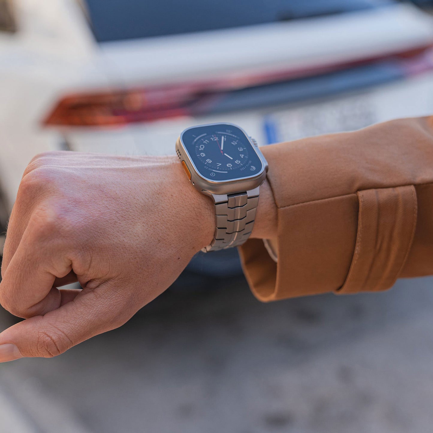 Eine Person in einer braunen Jacke präsentiert ein Arktisband Apple Watch TITAN Armband „Aviato Edition“ an ihrem Handgelenk, im Straßenhintergrund ist ein Teil eines weißen Autos zu sehen.