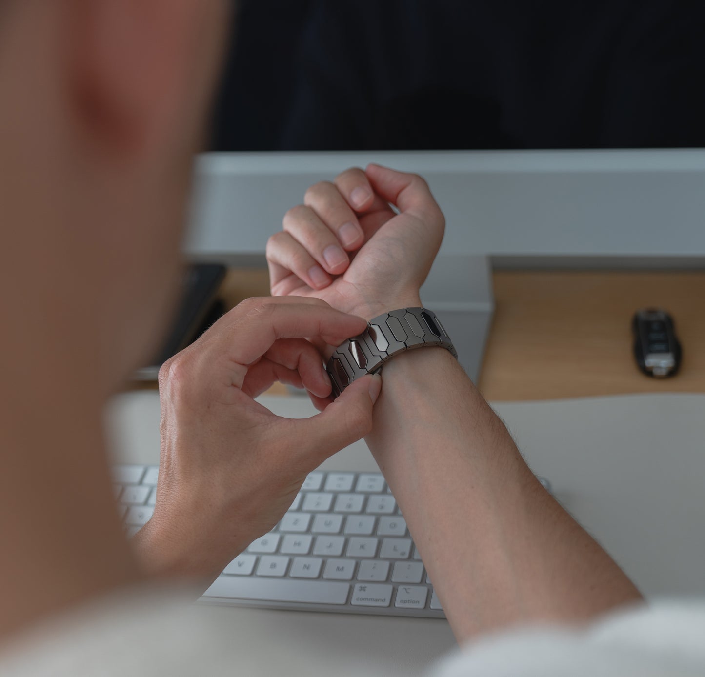 Eine Person passt ein Arktisband Apple Watch Ultra Titanarmband "Vectoron" mit hypoallergener DLC-Beschichtung an, während sie an einem Schreibtisch mit einer Tastatur und einem Monitor sitzt.