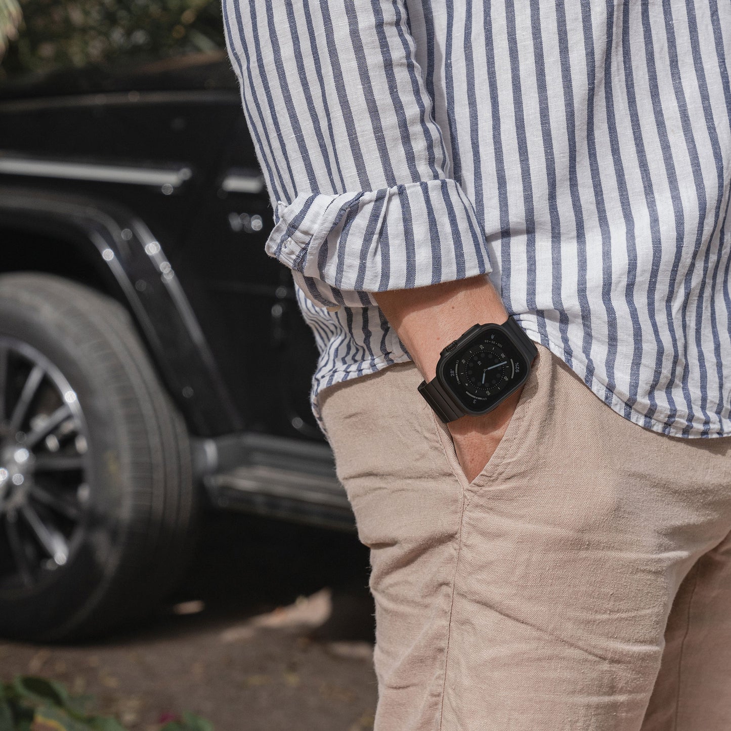 Eine Person in einem gestreiften Langarmhemd und einer beigen Hose steht mit einer Hand in der Tasche da und präsentiert ein Arktisband Apple Watch Ultra Titanarmband „Phantom“. Im Hintergrund sind das Rad und die Tür eines schwarzen Fahrzeugs zu sehen.