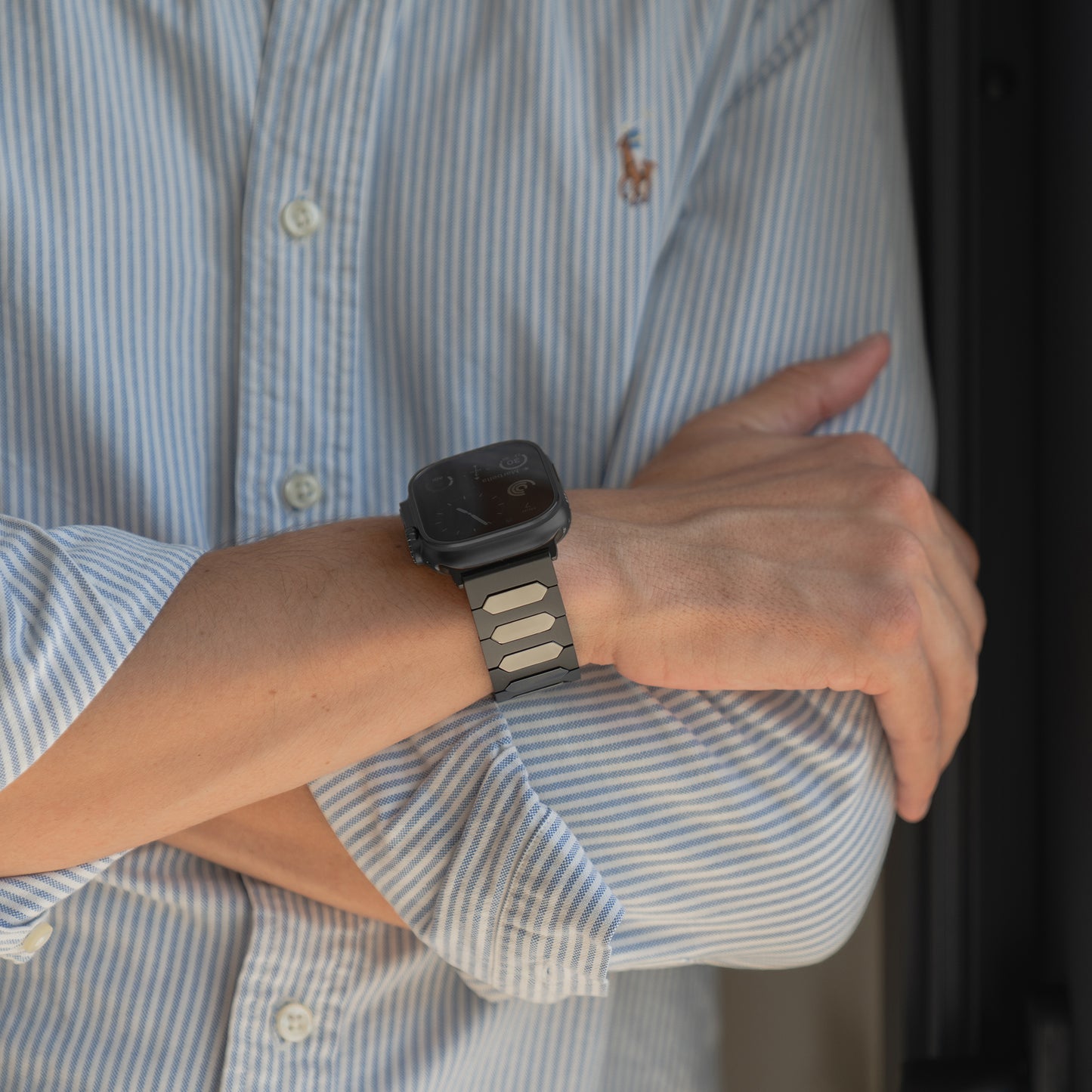 Person in einem blau gestreiften Hemd, die Arme verschränkt, trägt eine Arktisband Apple Watch Ultra Titanarmband "Vectoron" - eine schwarze Smartwatch mit einem silbernen Arktisband.