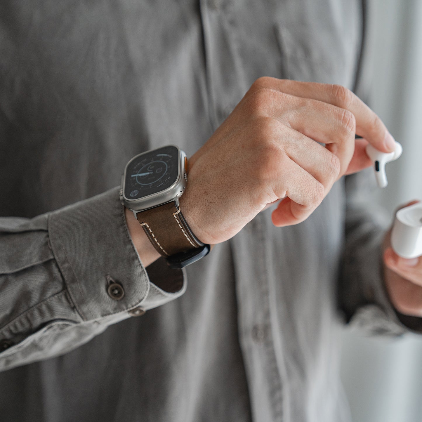 Eine Person trägt ein graues Hemd mit Knöpfen und ein Arktisband Apple Watch Ultra PALERMO GRANDE Lederarmband am linken Handgelenk und hält ein Paar kabellose Ohrhörer, wobei sie einen Ohrhörer in der Hand hält und in der anderen das Ladeetui.