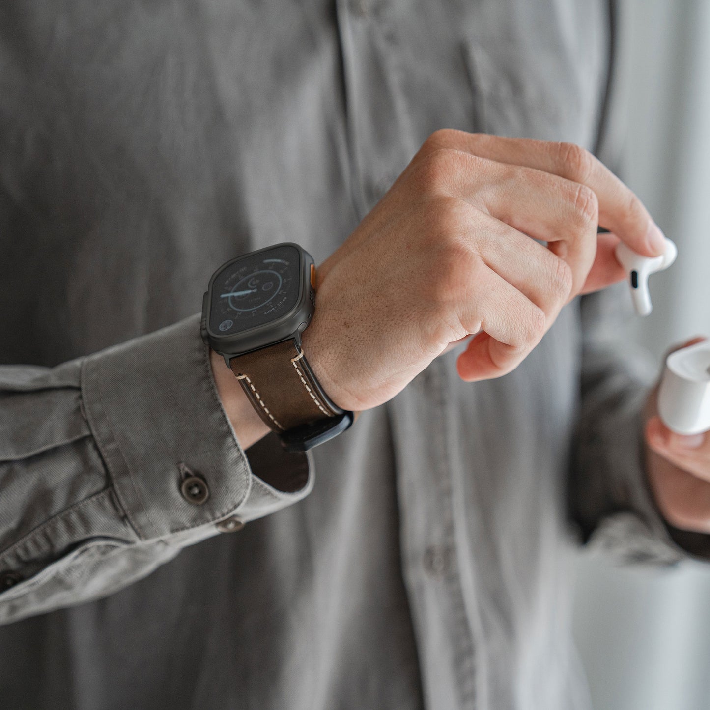 Eine Person in einem grauen Hemd trägt ein Arktisband Apple Watch Ultra PALERMO GRANDE Lederarmband am linken Handgelenk und hält kabellose Ohrhörer – einen Ohrhörer in der Hand, das Etui in der anderen.