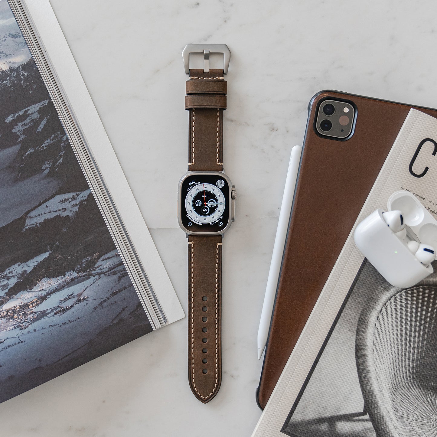 Ein Arktisband Apple Watch Ultra PALERMO GRANDE Lederarmband liegt auf einer Marmoroberfläche neben einem iPad mit einem Bleistift, einem offenen AirPods-Case und zwei Zeitschriften – eine mit Bergen, die andere mit einem Stuhl.