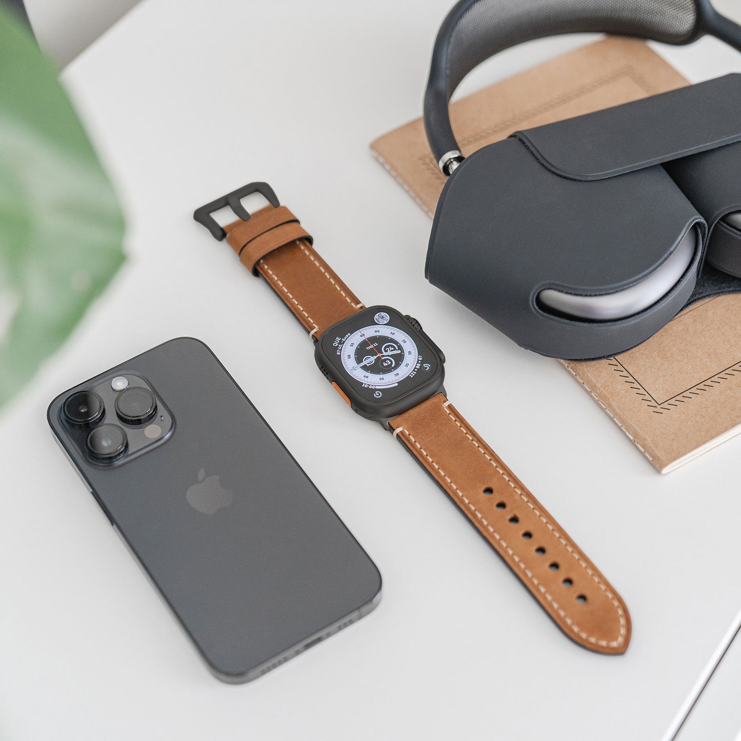 Ein schwarzes Smartphone, eine Apple Watch Ultra mit Arktisband und PALERMO GRANDE Lederarmband sowie schwarze Over-Ear-Kopfhörer liegen auf einer weißen Fläche neben einem hellbraunen Notebook.