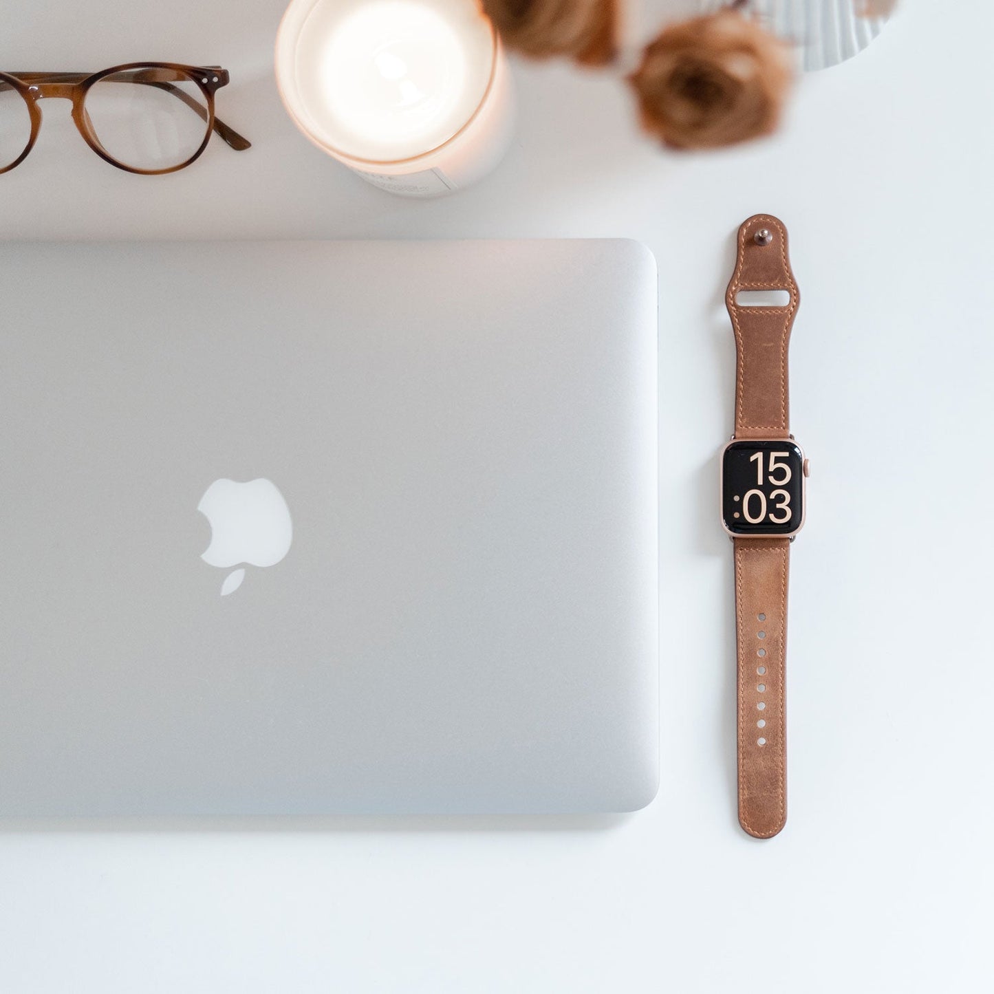 Ein geschlossenes silbernes MacBook, ein arktisband Apple Watch Lederarmband „Novus“ mit der Darstellung 15:03, eine Brille, eine brennende Kerze und eine Vase mit Trockenblumen schaffen einen minimalistischen Look auf einer weißen Oberfläche.