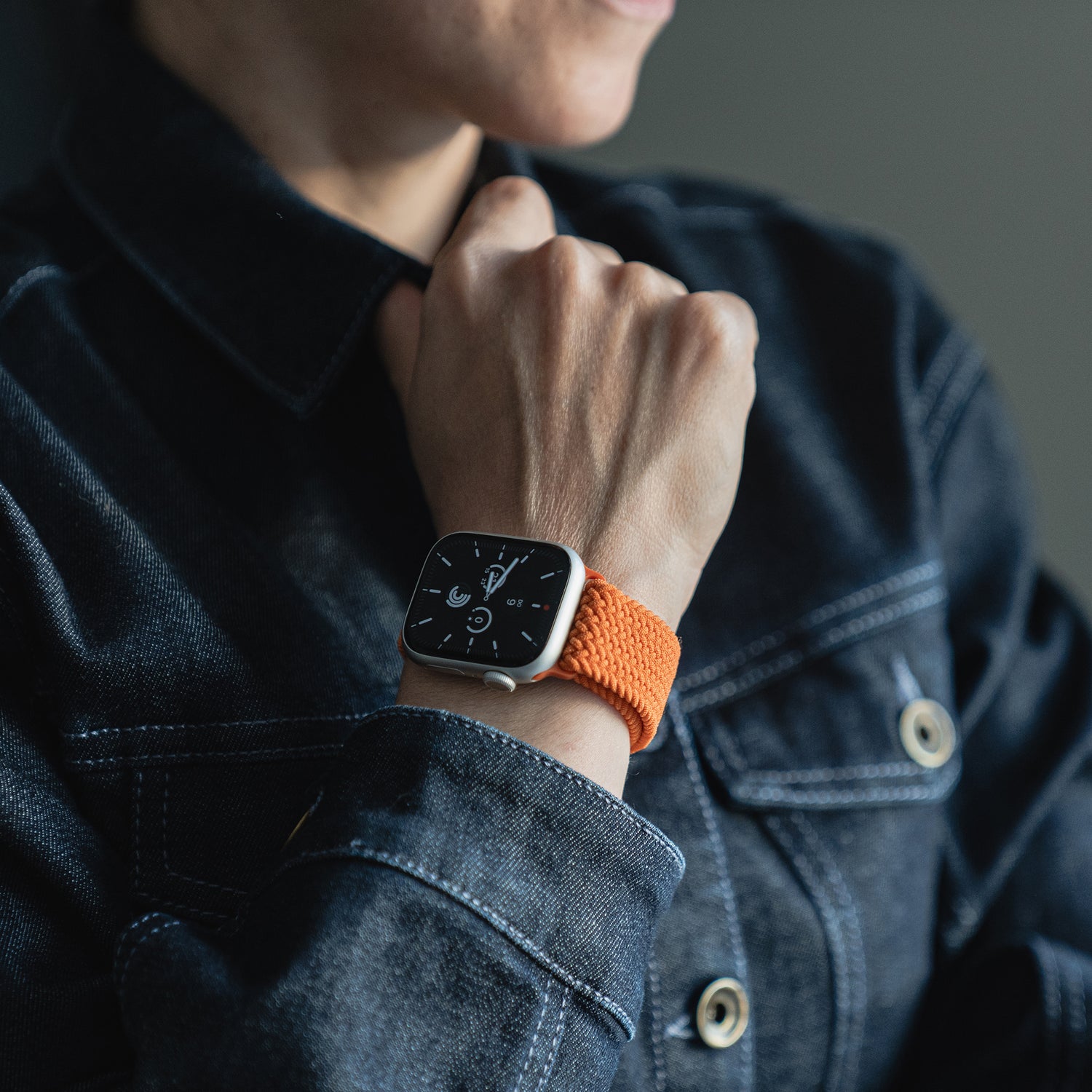Person, die eine dunkle Jeansjacke und eine Apple Watch mit dem arktisband Geflochtenen Apple Watch Armband „Casual“ mit Magnetverschluss von arktisband trägt, mit einem orangefarbenen geflochtenen Band am Handgelenk.