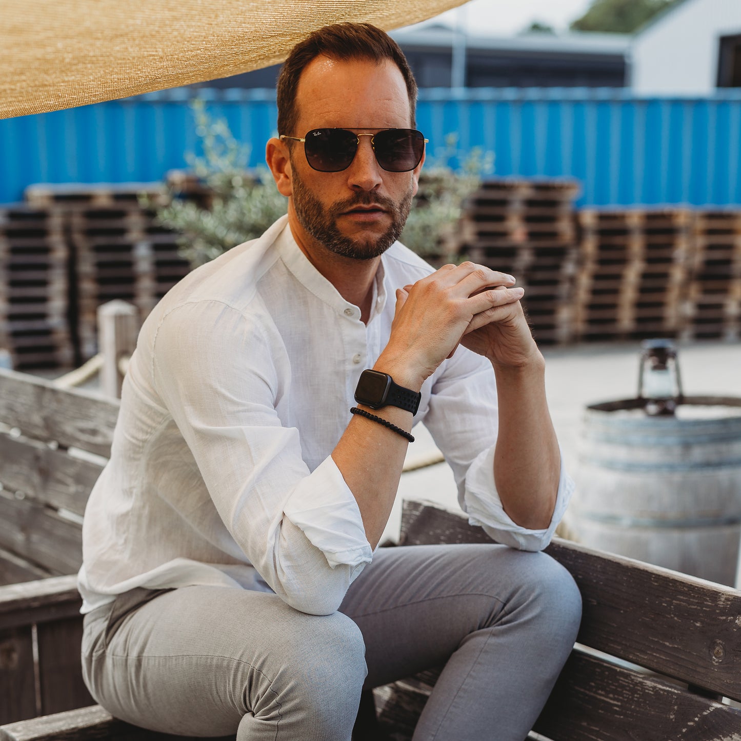 Ein Mann mit Sonnenbrille, weißem Hemd, grauer Hose und einem Arktisband Apple Watch Silikonarmband „Energy“ sitzt im Freien auf einer Holzbank, im Hintergrund sind gestapelte Paletten, ein Weinfass und ein blauer Schiffscontainer zu sehen.