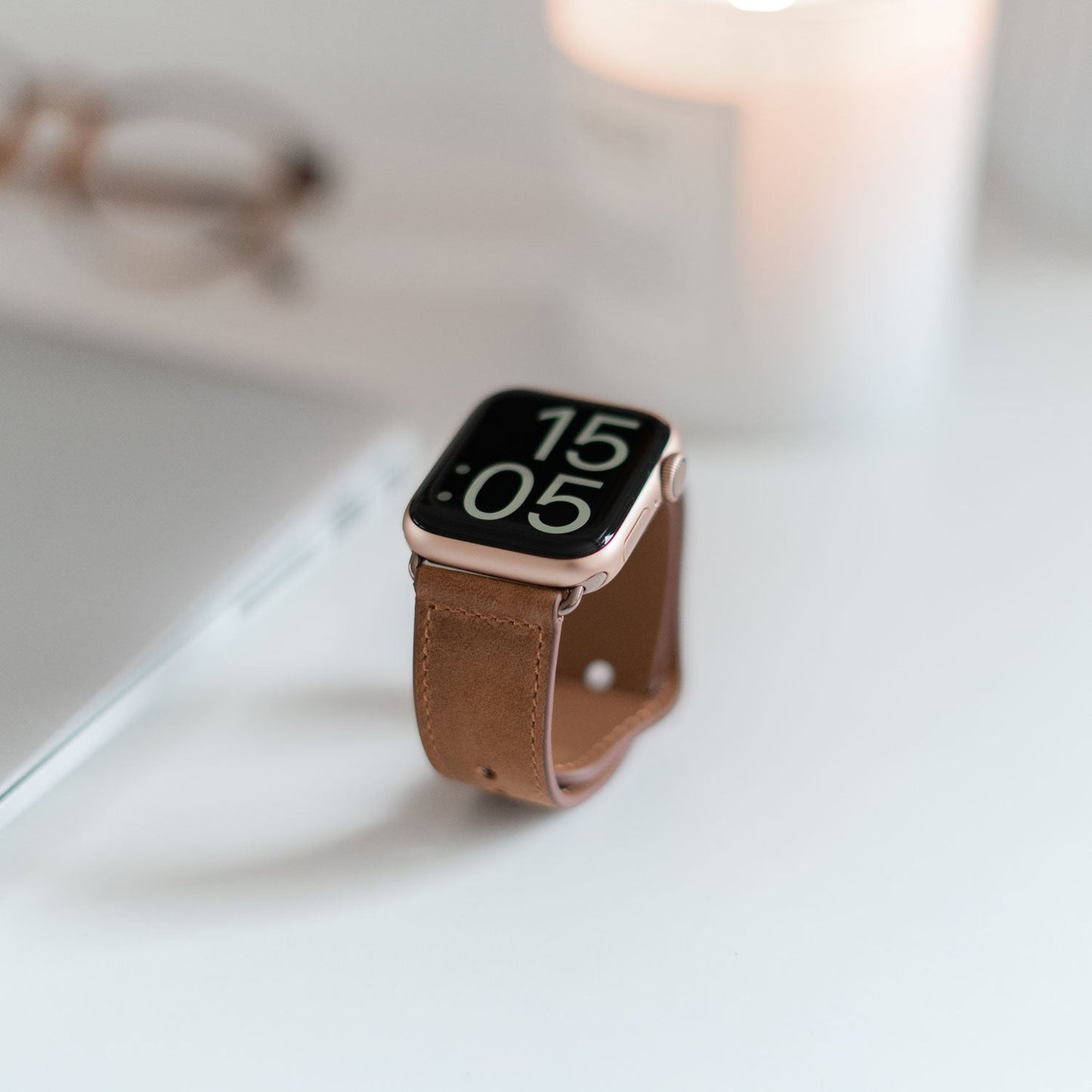 Eine Apple Watch mit dem arktisband Lederarmband „Novus“ von arktisband zeigt 15:05 Uhr. Im minimalistischen Stil liegt sie auf weißem Untergrund neben einem geschlossenen Laptop, einer Brille und einer brennenden Kerze.