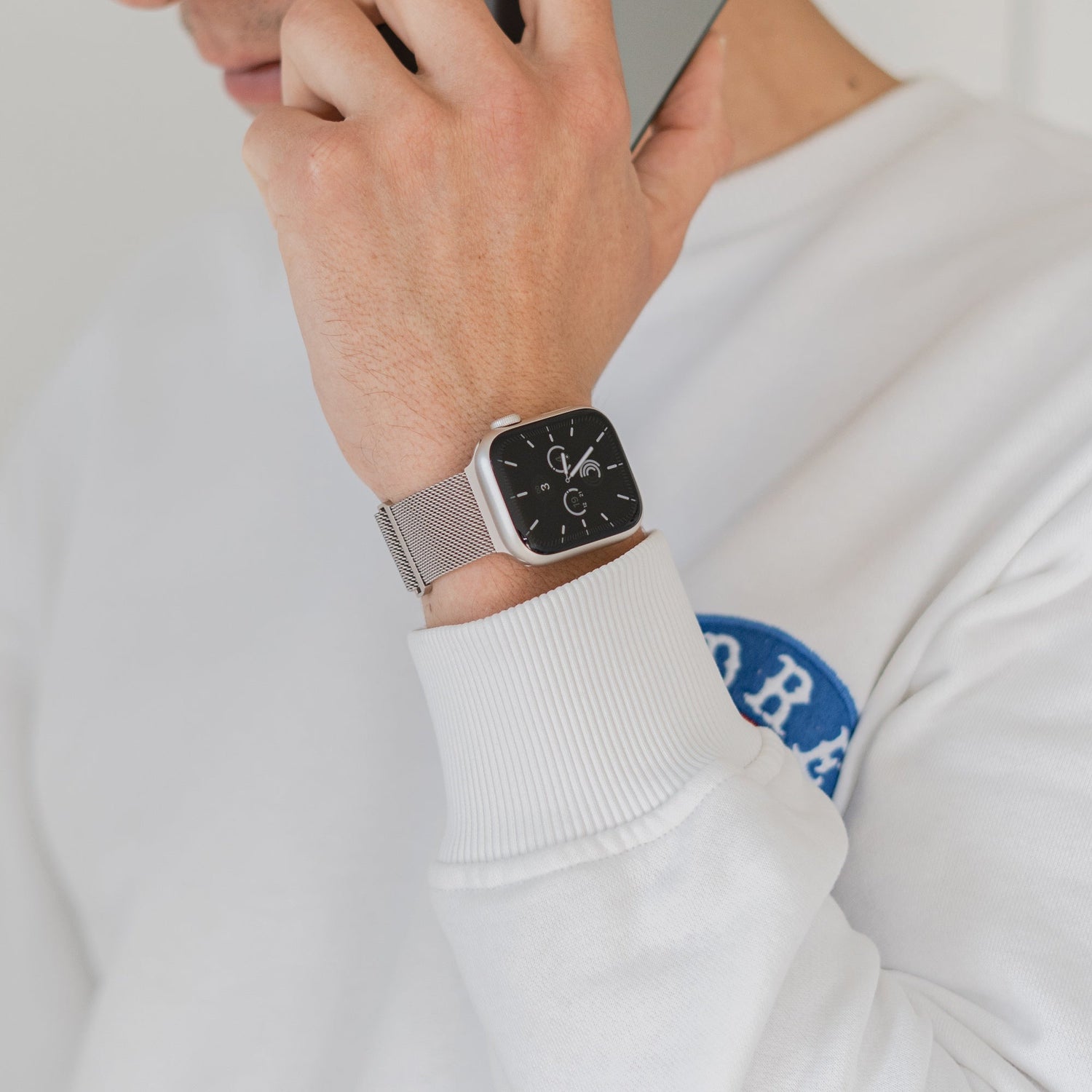 Eine Person in einem weißen Sweatshirt mit blauem Aufnäher telefoniert und trägt ein arktisband Apple Watch Milanaise Loop Armband von arktisband mit einem schwarzen quadratischen Zifferblatt am linken Handgelenk.