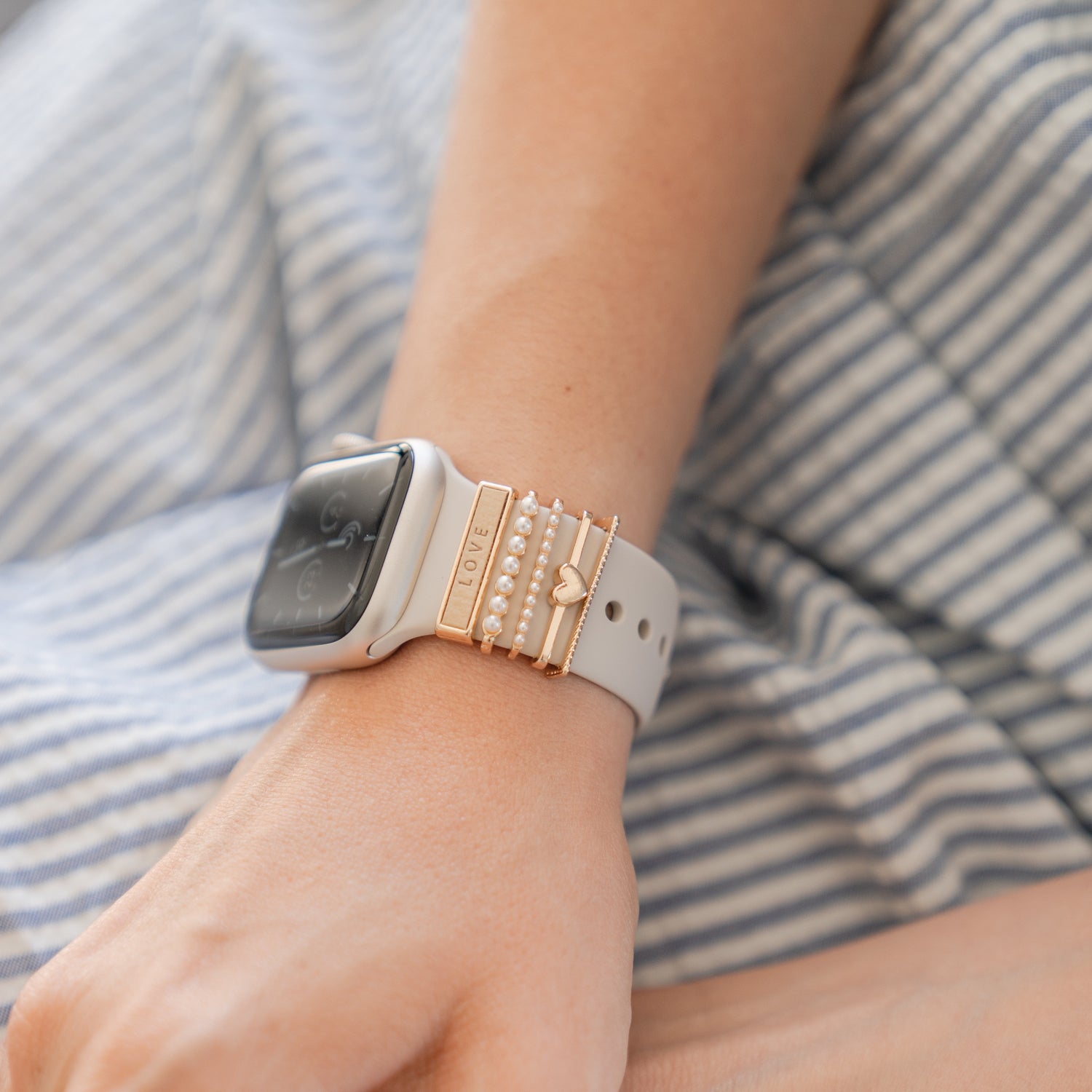 Eine Person trägt eine Arktisband Apple Watch mit goldenen „Love“-Anhängern von Arktisband und sitzt auf blauen und weißen Streifen – und präsentiert ein schickes Accessoire.