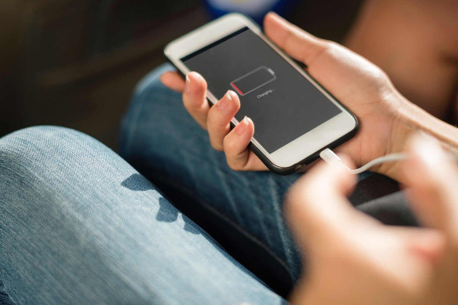 Eine Person hält ein Telefon mit einem Symbol für niedrigen Akkustand und lädt es auf, während sie in Jeans sitzt.