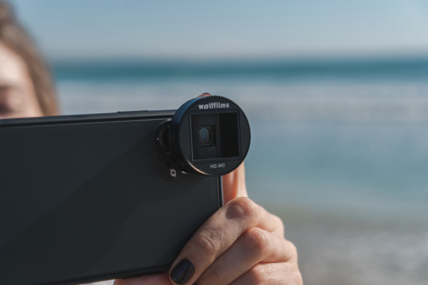 Eine Person hält ein Smartphone mit einem Wolffilms-Objektivaufsatz am Meer und macht ein Foto.