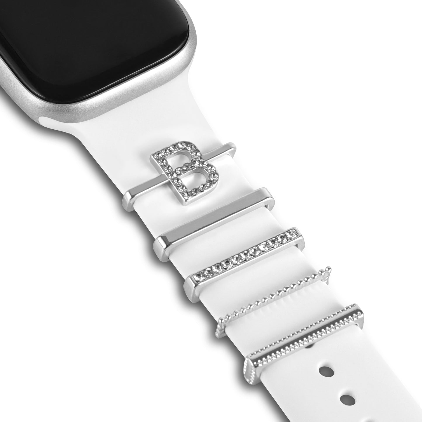 Eine Nahaufnahme der arktisband Apple Watch Charms „B Style“ von arktisband zeigt ein weißes Band mit vier dekorativen Charms aus Edelstahl und Glas, darunter ein mit Edelsteinen besetztes „B“; das silberne Smartwatch-Gehäuse hat ein schwarzes Display.