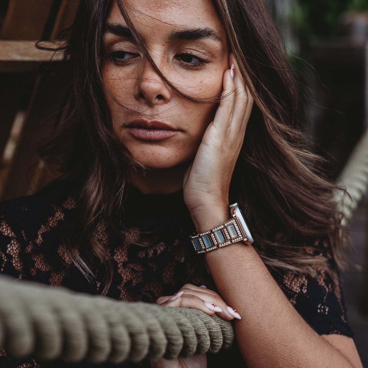 Eine Frau mit langen braunen Haaren stützt ihr Gesicht auf ihre Hand und blickt neutral nach unten. Sie trägt ein schwarzes Spitzentop und das Arktisband Apple Watch Gliederarmband „Senorita“. Im Vordergrund sind dicke Seile zu sehen.