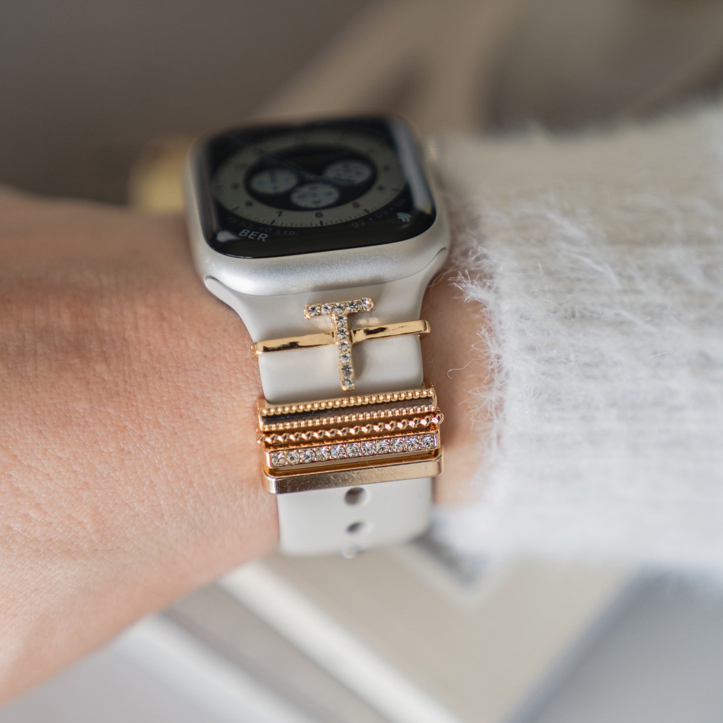 Eine Nahaufnahme eines Handgelenks mit einer Arktisband Apple Watch mit weißem Armband, verziert mit Arktisband-Anhängern im „T-Stil“. Die Person trägt außerdem einen flauschigen weißen Pullover.