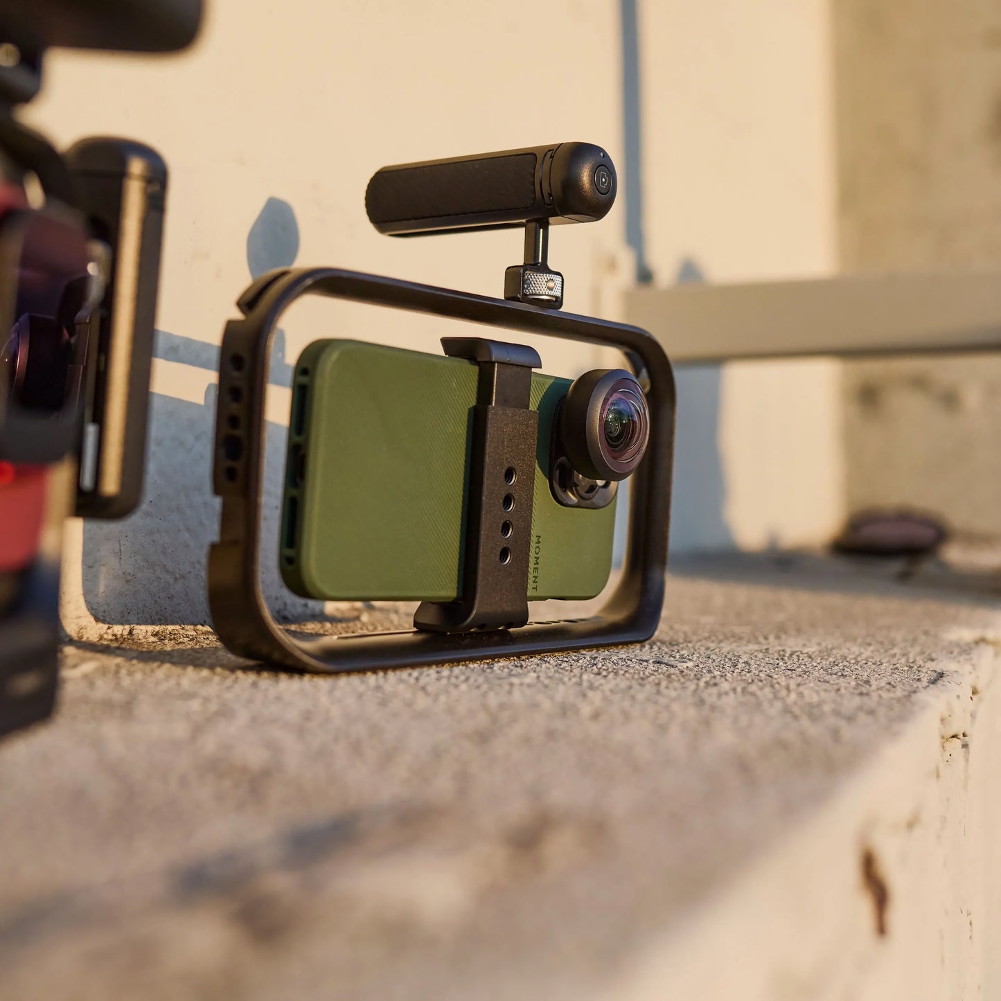 Der MOMENT Basecage - Fast and Light Mobile Cage hält ein Smartphone mit externem Objektiv und Mikrofon, das auf einer Betonfläche im Sonnenlicht aufgestellt wird.