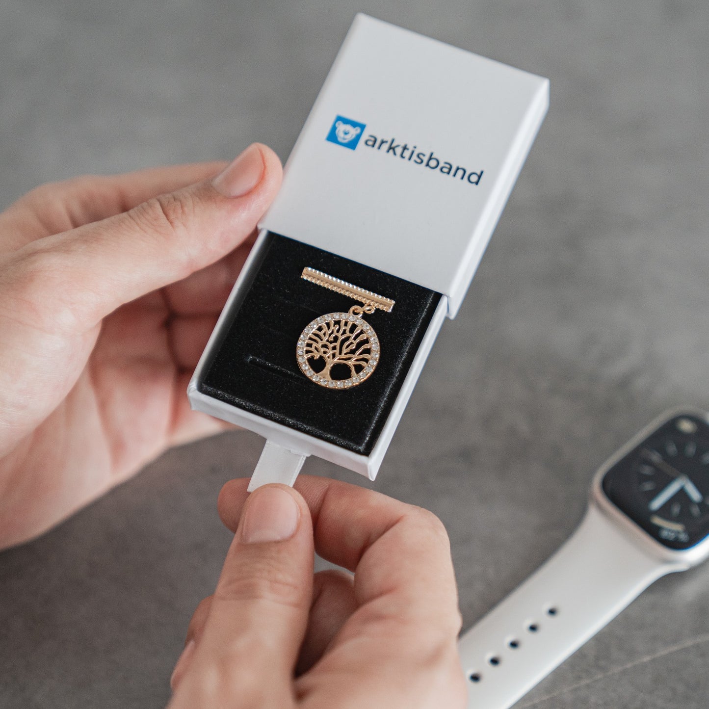 Eine Person öffnet eine Arktisband-Box und findet darin einen goldenen Lebensbaum-Anhänger. Mit Arktisband-Apple-Watch-Armbändern zeigt sie, wie sie ihren Stil mit diesen einzigartigen Accessoires von Arktisband personalisieren kann.