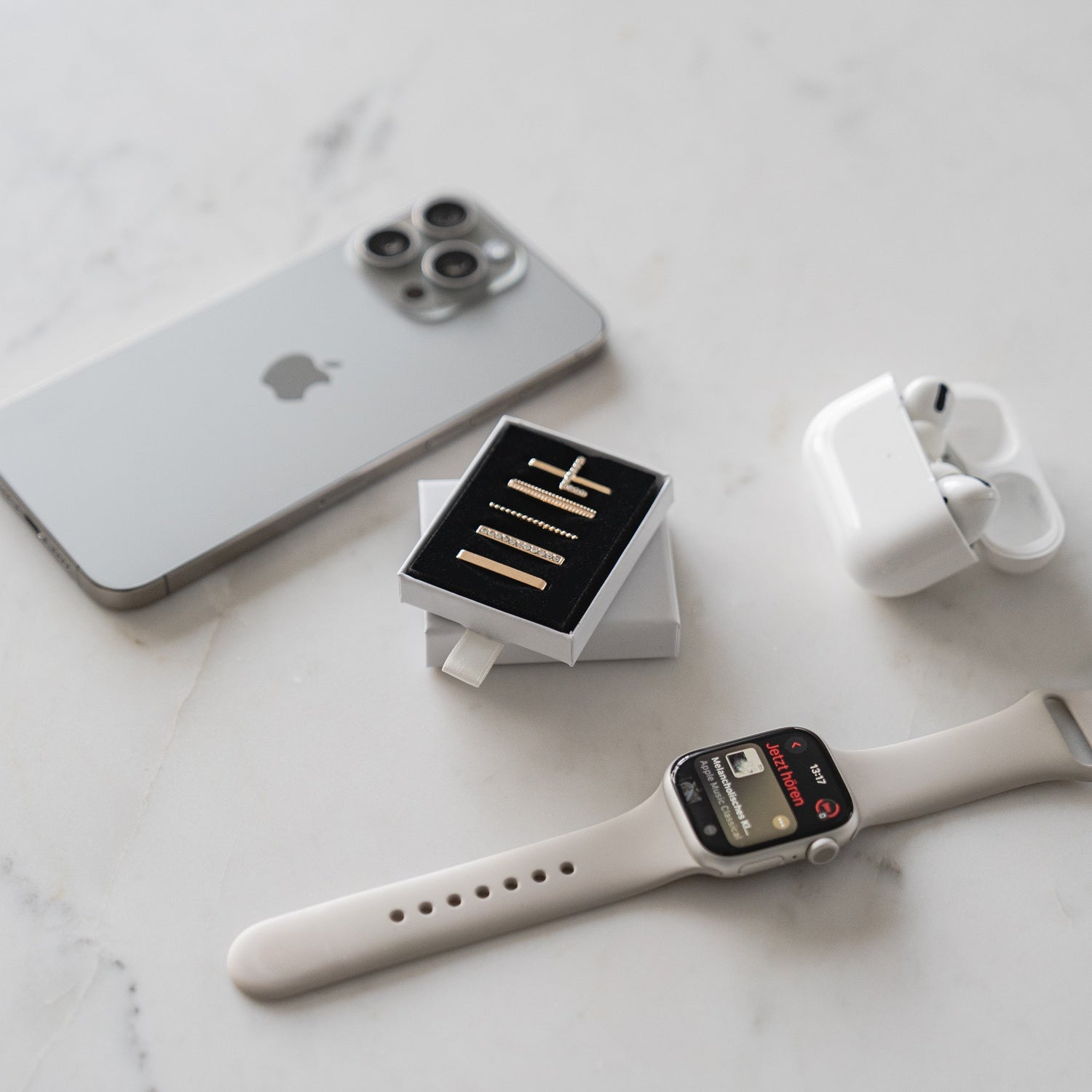 Ein silbernes iPhone, weiße kabellose Ohrhörer in einem Ladeetui, eine weiße Smartwatch mit Arktisband Apple Watch Charms „L Style“ und eine kleine Schachtel mit goldenen Manschettenknöpfen sind auf einer hellen Oberfläche angeordnet.
