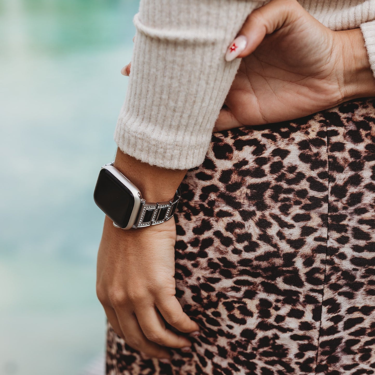 Eine Person mit einer Hand auf der Hüfte trägt einen cremefarbenen Rippenpullover, einen Rock mit Leopardenmuster und ein arktisband Apple Watch Gliederarmband „Senorita“ von arktisband. Ihr Nageldesign zeigt einen kleinen roten Stern auf einem Fingernagel.