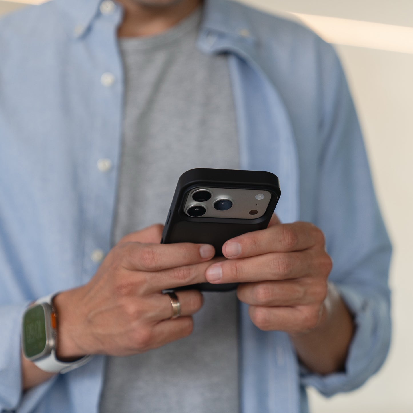 Eine Person in einem blauen Hemd benutzt ein Smartphone mit dem Arktis iPhone 17 Pro Solid Case, während sie eine Smartwatch und einen Ehering trägt.
