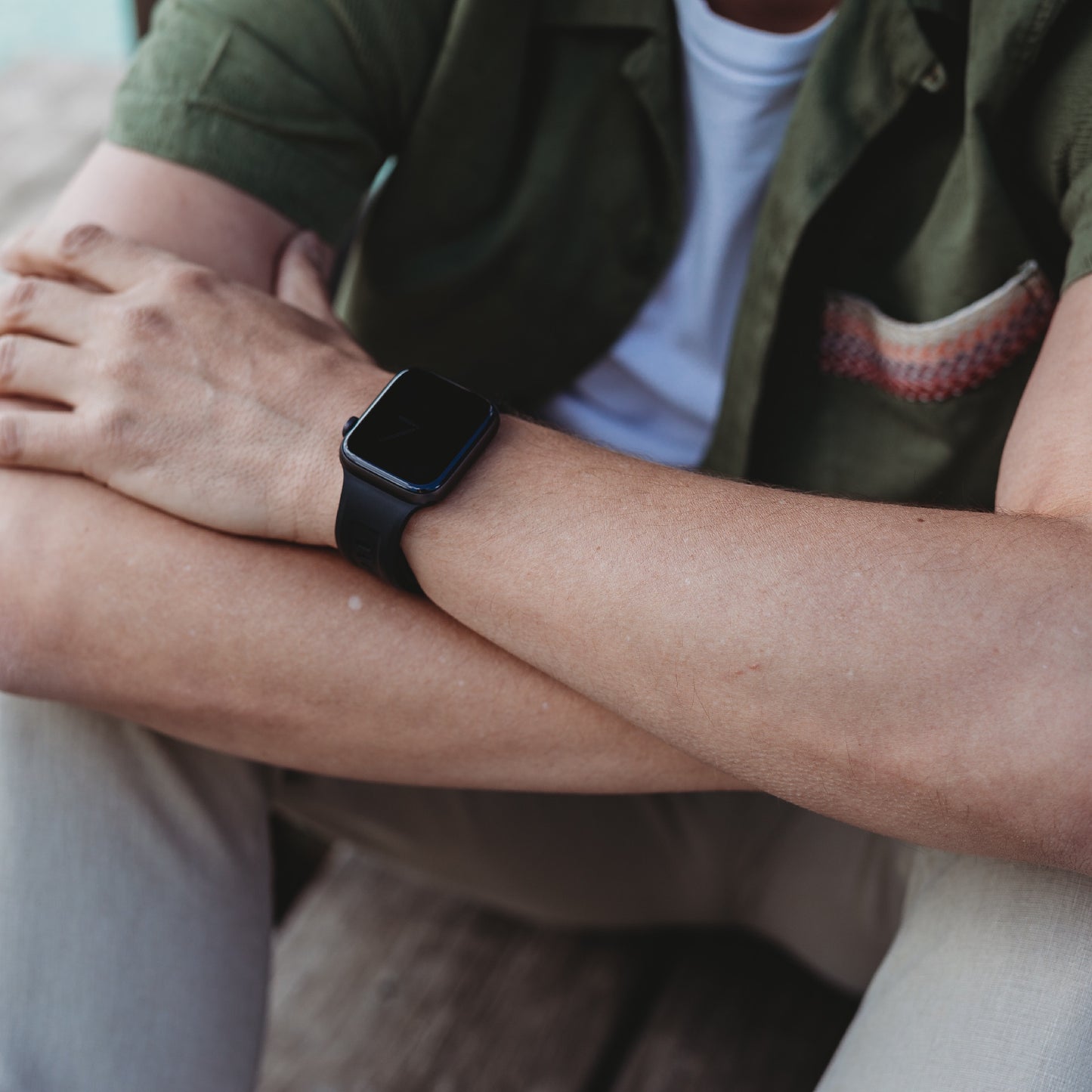 Eine Person trägt das arktisband Apple Watch FKM Sportarmband „Sweat“ (dunkel), sitzt mit verschränkten Armen im Freien, bekleidet mit einem grünen Hemd mit gemusterter Tasche und einer hellen Hose; auf dem Bild sind Oberkörper und Arm zu sehen.
