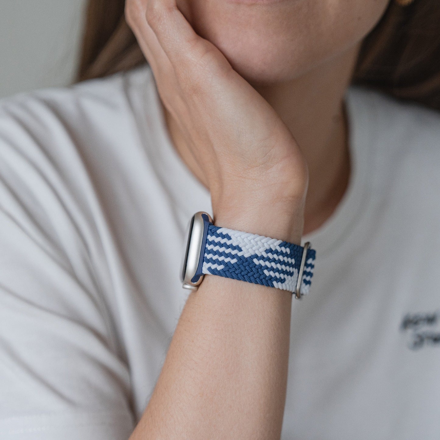 Eine Person in einem weißen Hemd stützt ihr Kinn auf die Hand und präsentiert ein geflochtenes Flex Loop TwoTone-Armband für die Apple Watch mit einem stilvollen Edelstahlverschluss an ihrem Handgelenk.