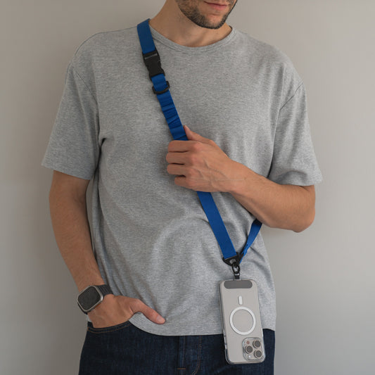 Eine Person in einem grauen T-Shirt und Jeans steht an einer hellen Wand. Sie trägt ein arktis Smartphone-Gurtband-Lanyard, das an ihrer Telefonhülle befestigt ist, quer über ihrem Körper und eine Smartwatch am linken Handgelenk.