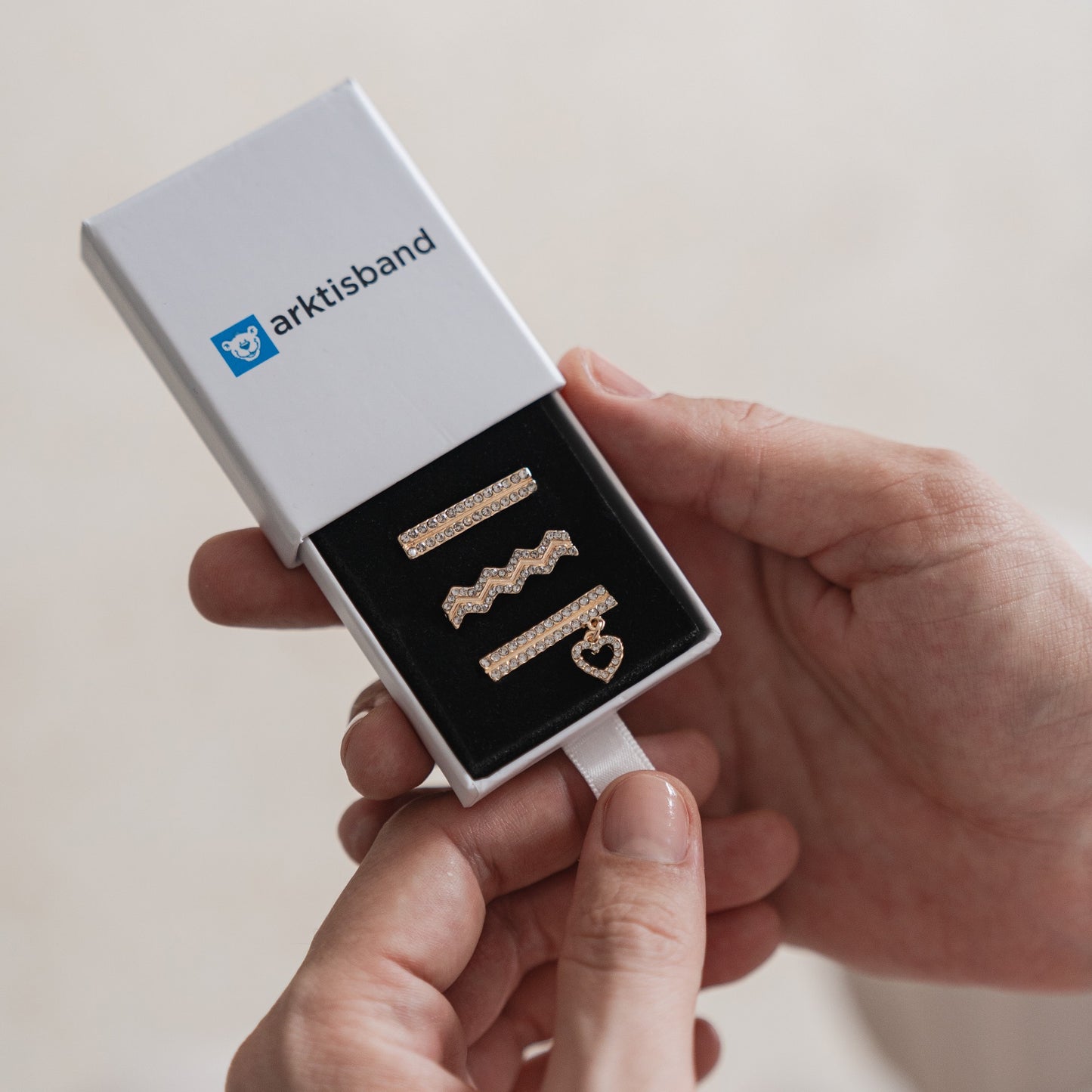 Eine Person hält eine offene weiße Arktisband-Box mit drei goldenen Haarspangen – gerade, zickzackförmig und herzförmig – ideal zum Personalisieren oder Kombinieren mit Ihren Arktisband-Apple-Watch-Armband-Charms.