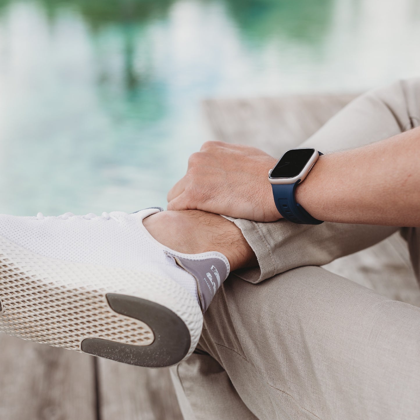 Eine Person in heller Hose, weißen Turnschuhen und einem blauen Arktisband Apple Watch FKM Sportarmband „Sweat“ sitzt im Schneidersitz auf einem Holzdeck am Wasser, wobei nur die Unterschenkel, Hände und ein Teil des Arms sichtbar sind.