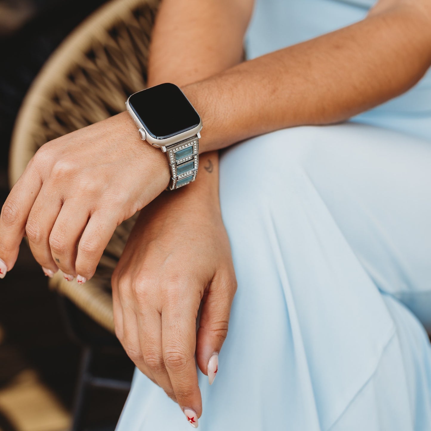Eine Person, die das Arktisband Apple Watch Gliederarmband „Senorita“ mit Edelstahlarmband und Strasssteinenbesatz trägt, sitzt mit verschränkten Armen über einer hellblauen Hose und hat weiße Nägel mit kleinen roten Mustern zur Schau gestellt.