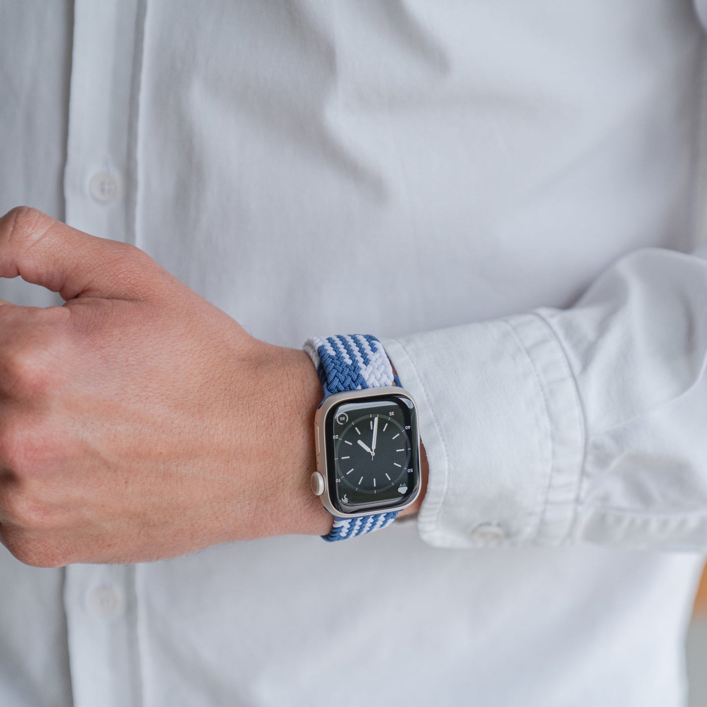 Eine Person in einem weißen Hemd mit Knöpfen ist von den Schultern abwärts zu sehen, wie sie ihre Ärmel zurechtrückt, während sie ein geflochtenes Flex Loop TwoTone-Armband mit Arktisband für die Apple Watch mit schwarzem Zifferblatt und Edelstahlverschluss trägt.