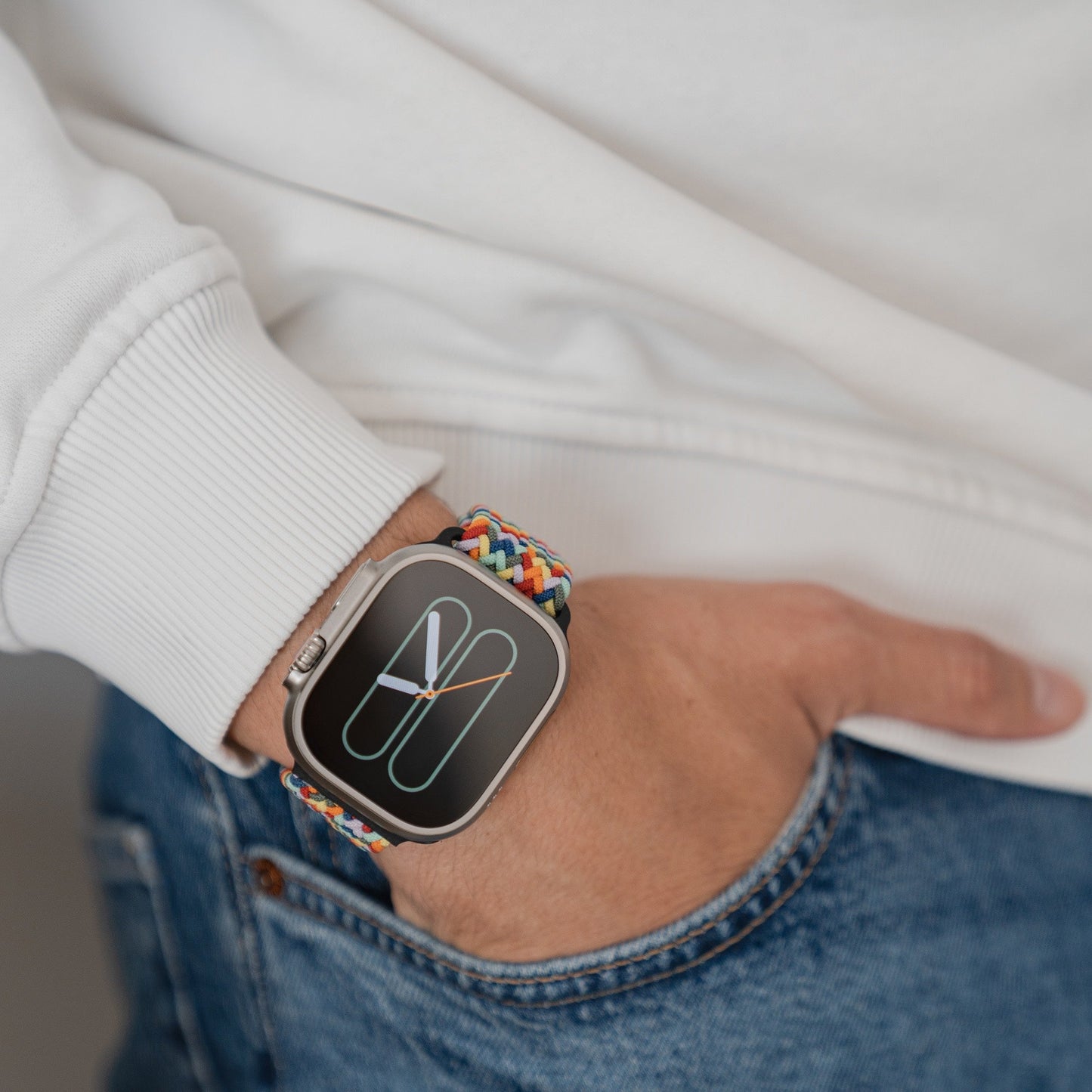Eine Person in weißem Sweatshirt und Blue Jeans, die linke Hand in der Hosentasche, trägt eine arktisband Apple Watch mit dem arktisband geflochtenen Flex Loop Multicolor Armband. Die Uhr zeigt ein minimalistisches Zifferblatt.